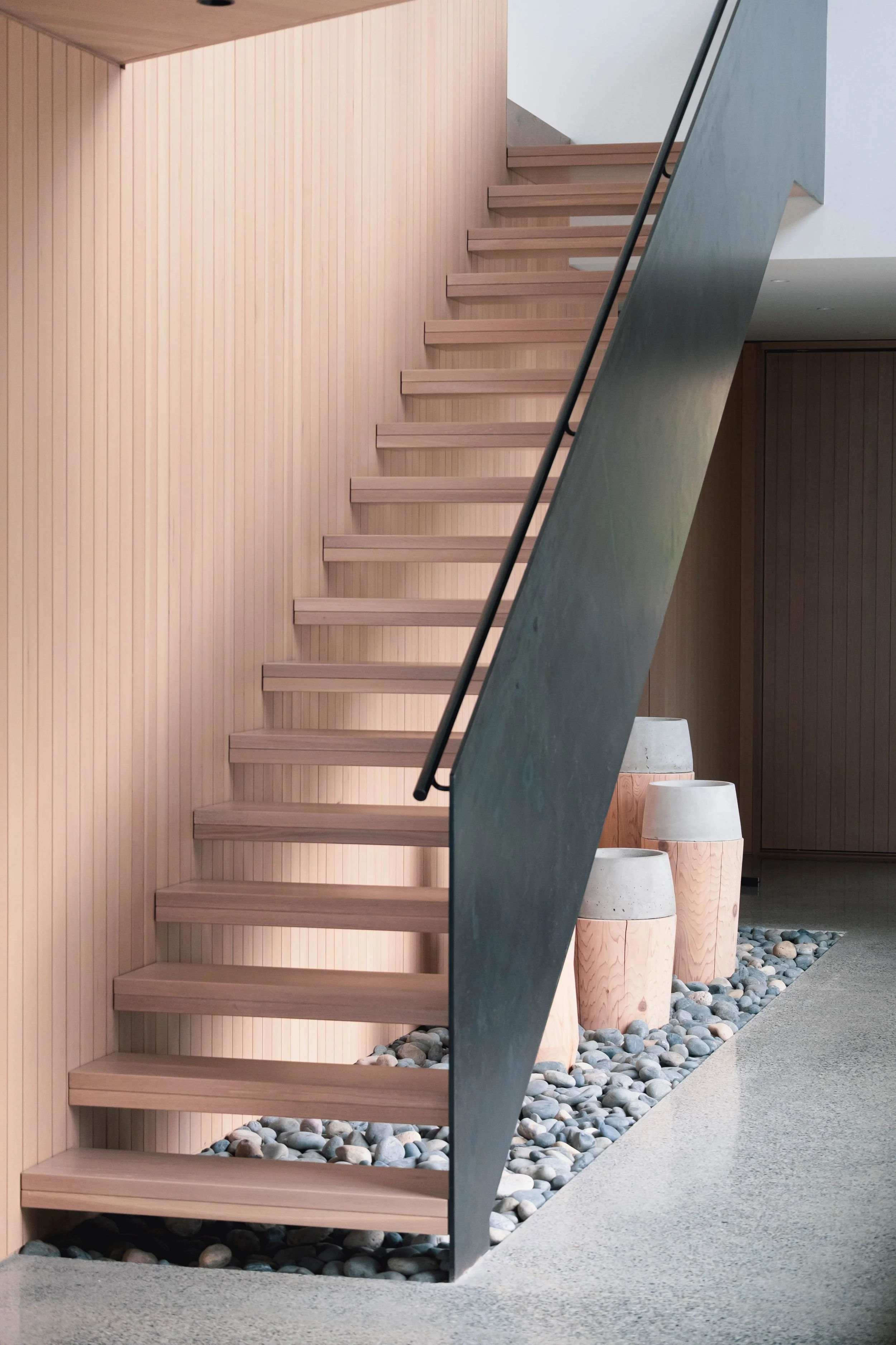 Modern indoor staircase with wooden steps, black handrail, and a pebble-filled area with three large, textured planters.