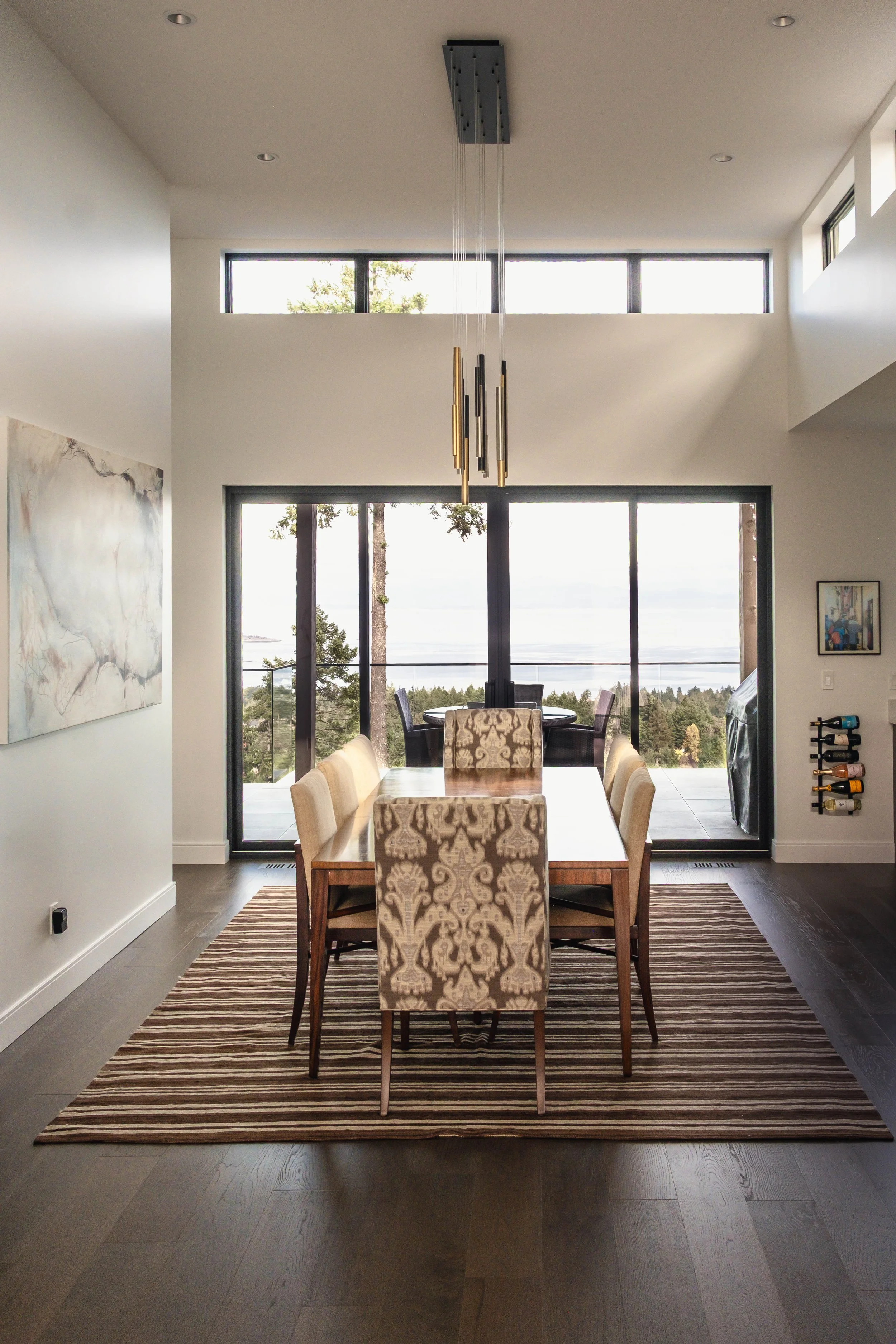 Modern dining room with a wooden table, upholstered chairs, large glass sliding doors leading to a balcony with outdoor seating, and a view of trees and water in the background.
