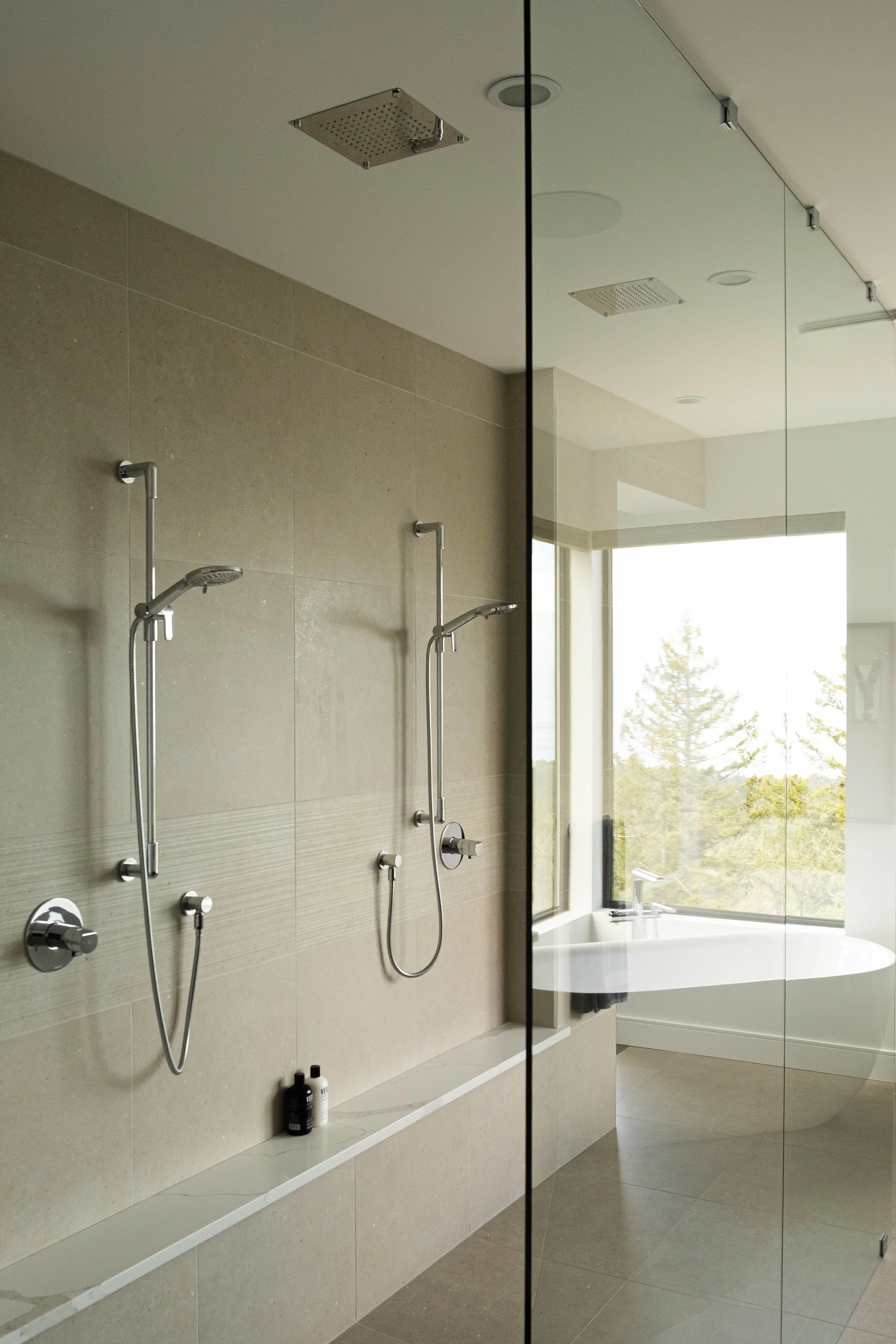 Modern bathroom with dual showers and a bathtub near a large window with a view of trees.