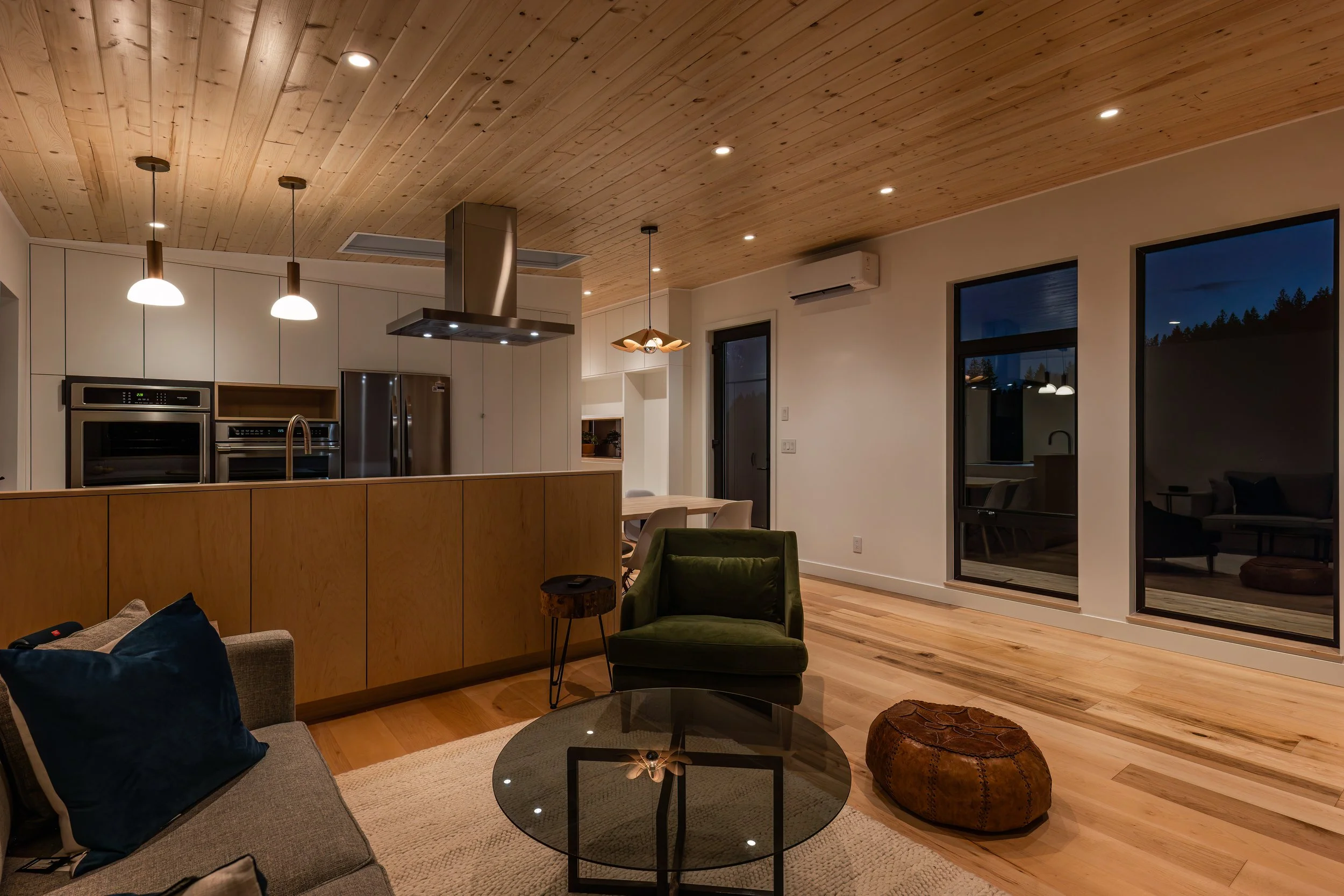 Interior view of a modern living room with wood ceiling and floor, large windows, and a kitchen in the background. Includes a sofa with pillows, a green armchair, a glass coffee table, and a brown pouf.