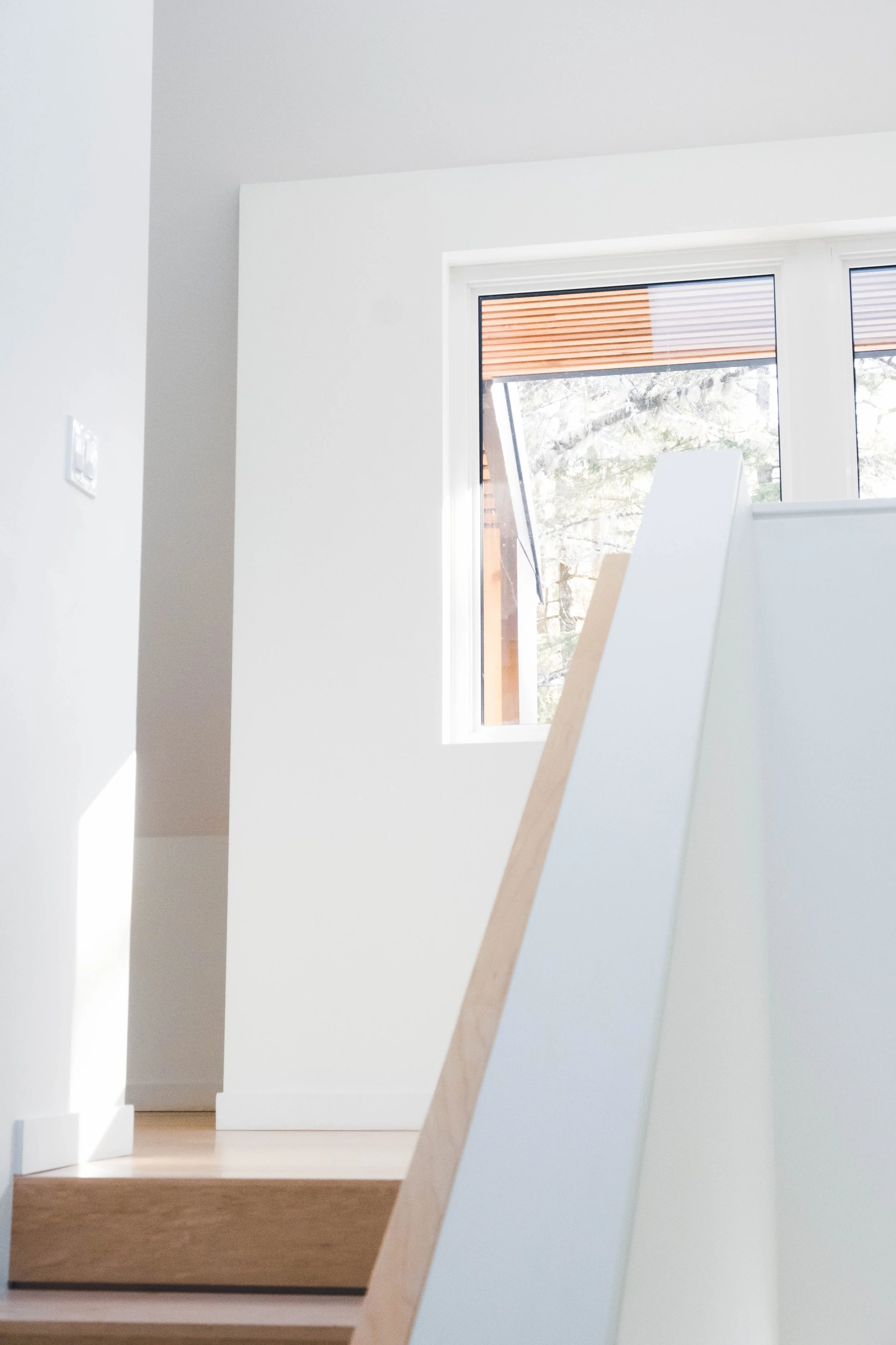 Interior view of a minimalist room with white walls, a wooden staircase, and a large window letting in natural light, overlooking trees outside.