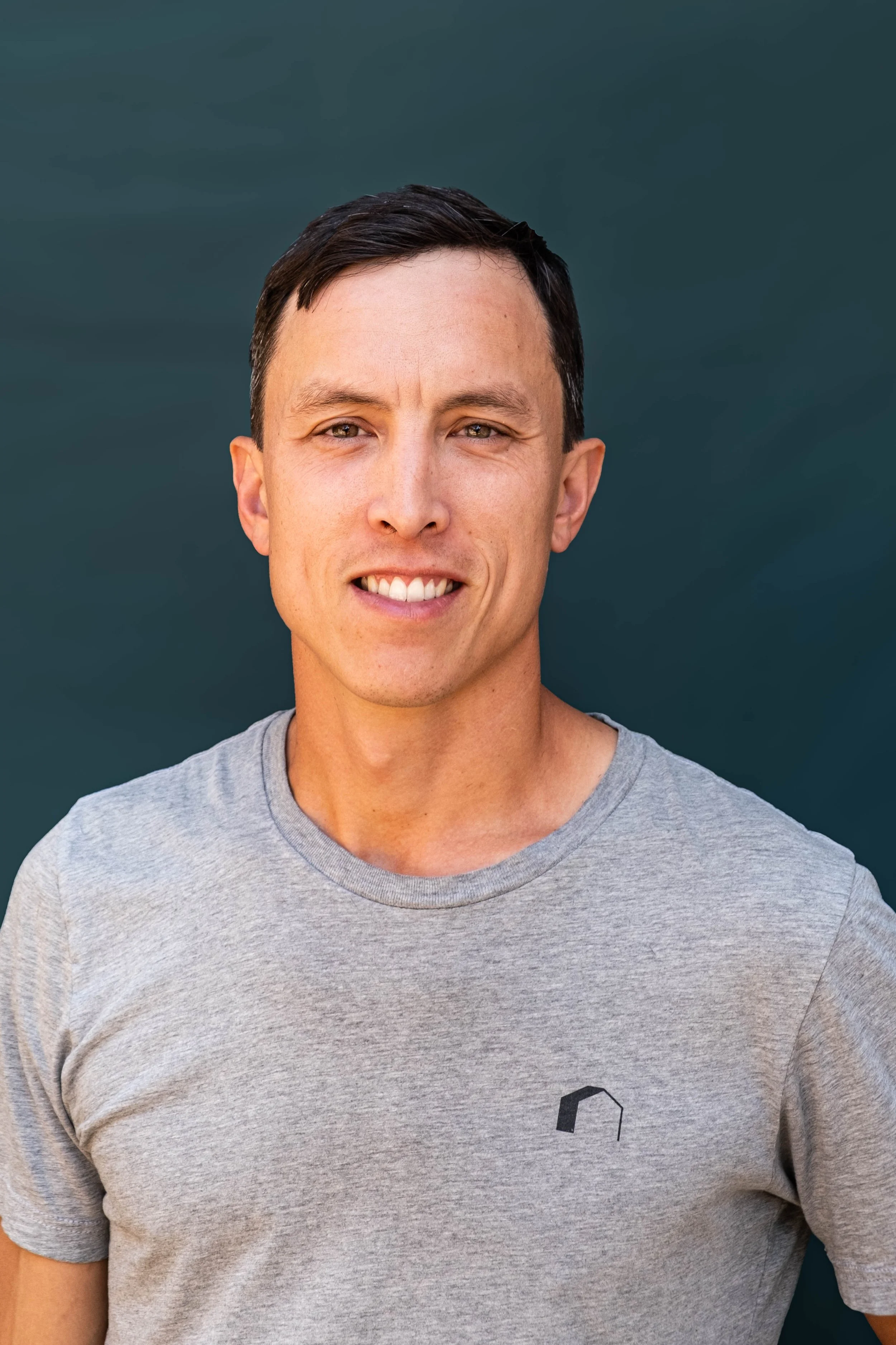 A man smiling outdoors against a dark background, wearing a gray t-shirt with a small logo.