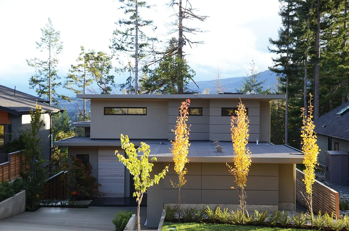 Modern two-story house with large windows, surrounded by trees with autumn foliage, set in a forested area with mountains in the background.