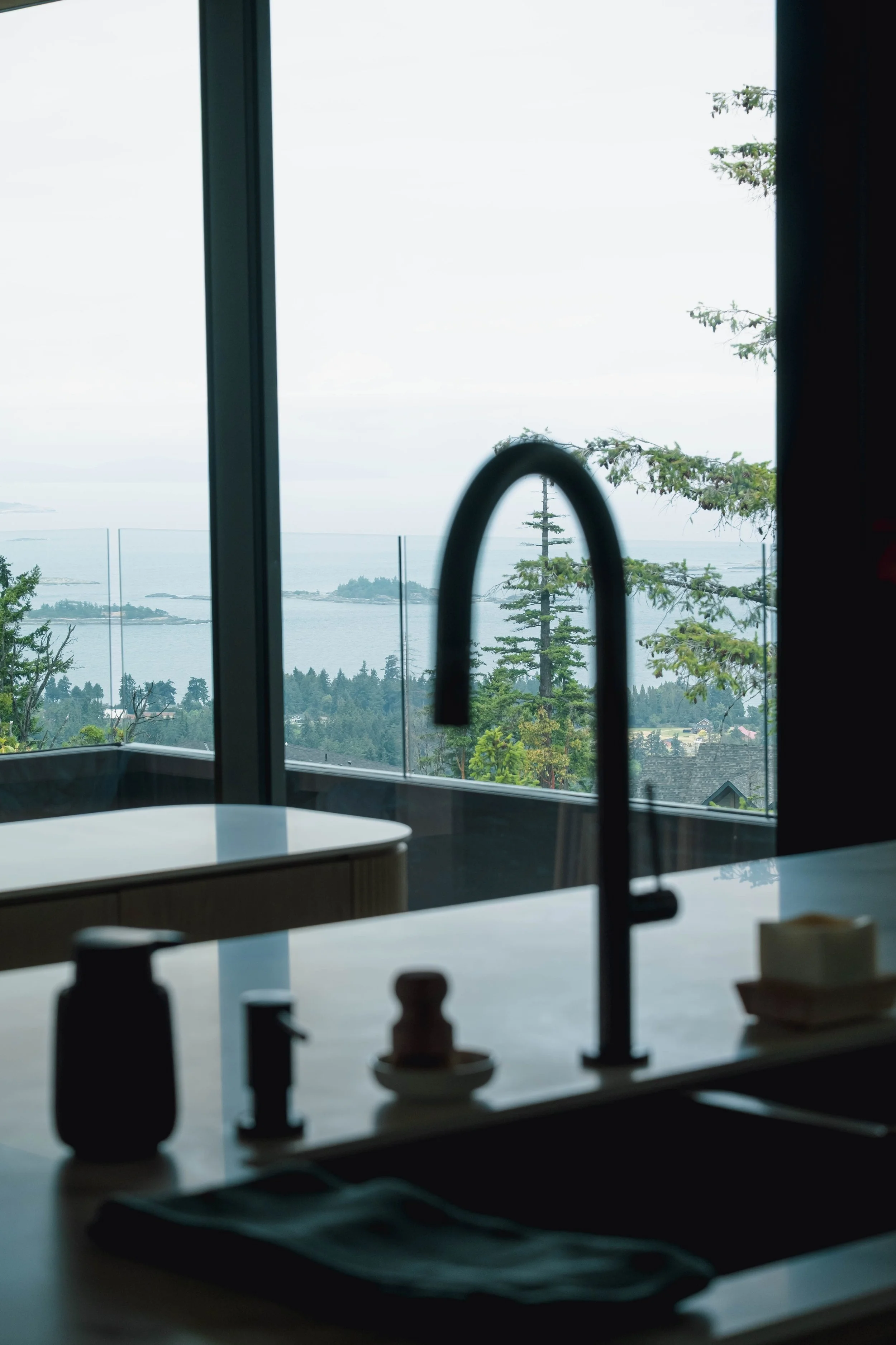 A kitchen countertop with a modern black faucet, soap dispenser, and a stack of yellow soap bars, with a large window showing a view of trees, the ocean, and an overcast sky.