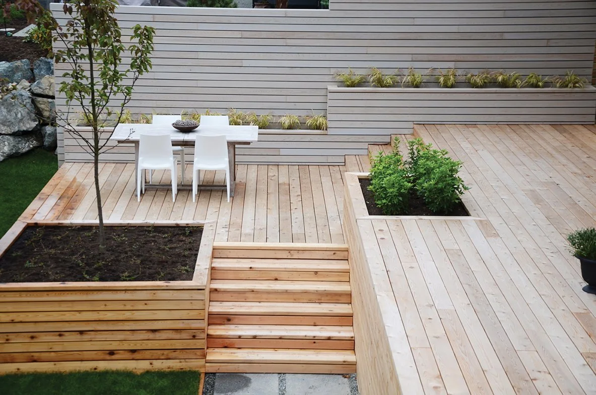 A modern outdoor patio with wooden decking, built-in wooden planters with soil and plants, a small table with four white chairs, and some greenery and rocks in the background.