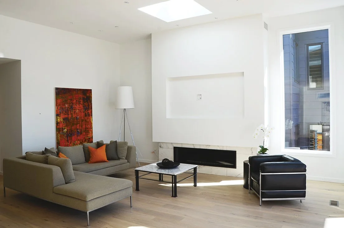 Minimalist living room with a beige sectional sofa, an orange throw pillow, a white floor lamp, a small marble-top coffee table, a black armchair, a large window, and a red abstract painting on the wall.