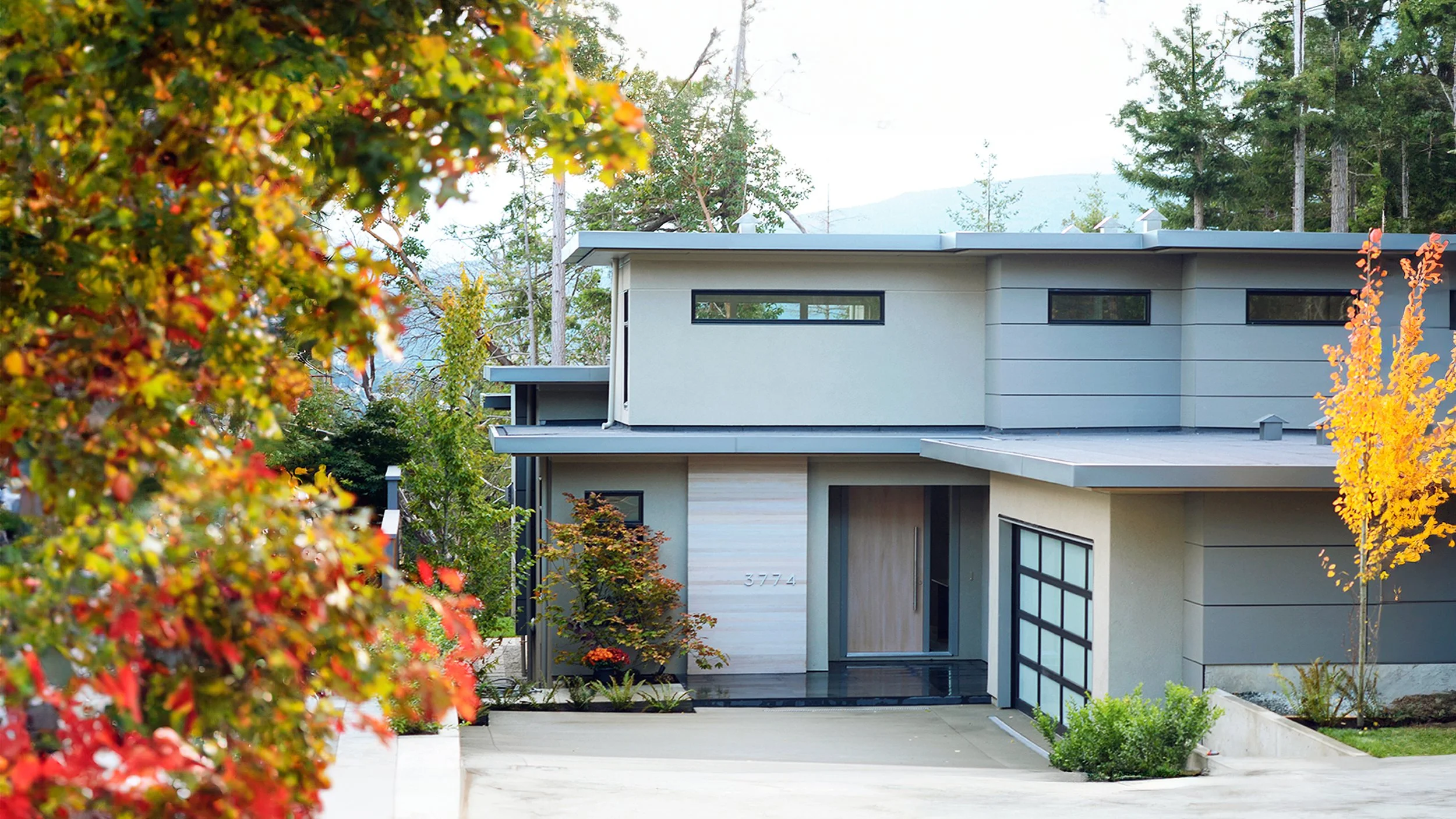 Modern house with gray exterior, large garage door, and surrounding trees with fall foliage.