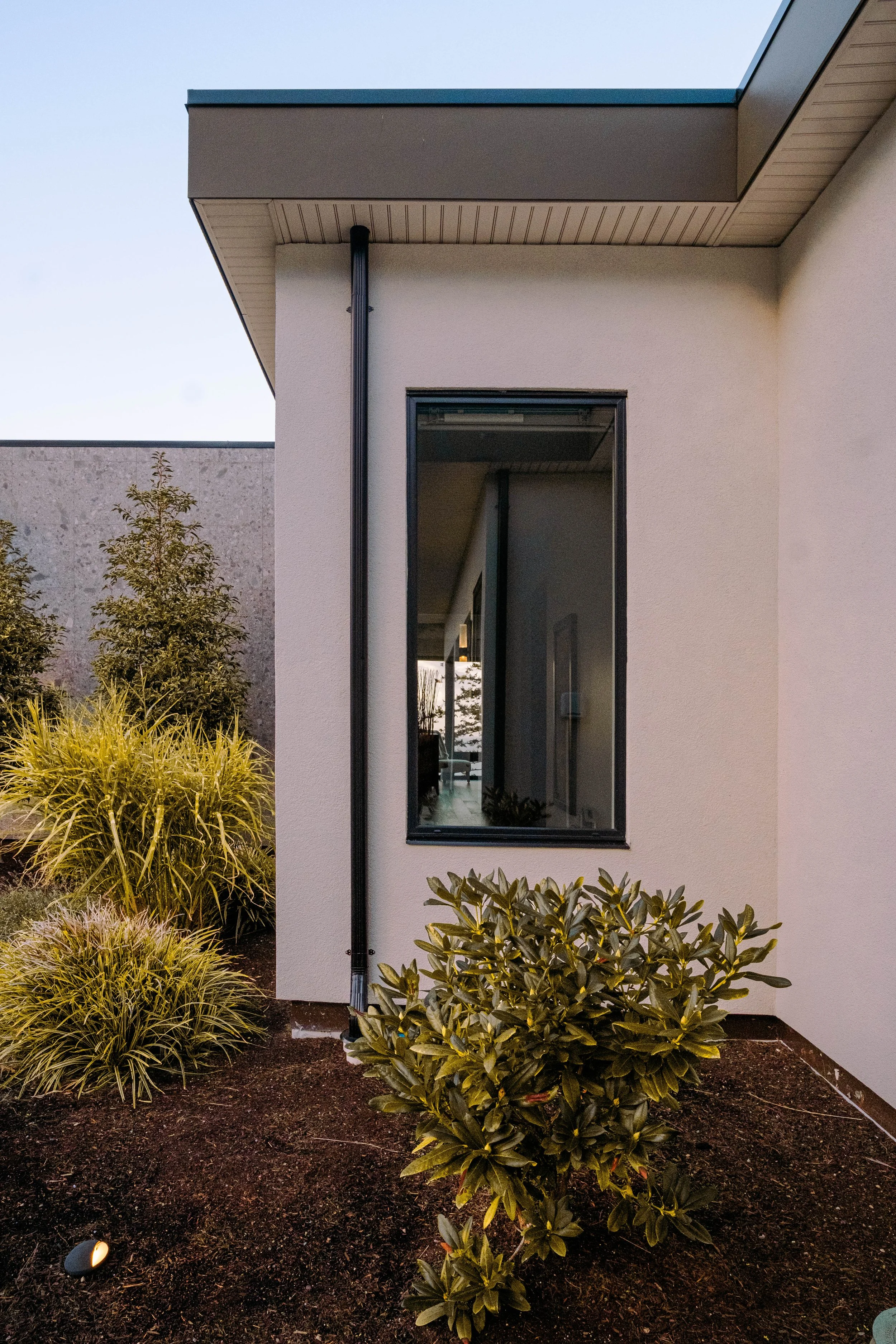 A modern house exterior with a beige stucco wall, a tall narrow window, and landscaped bushes. A black downspout runs along the wall, and the sky is visible above the roof.