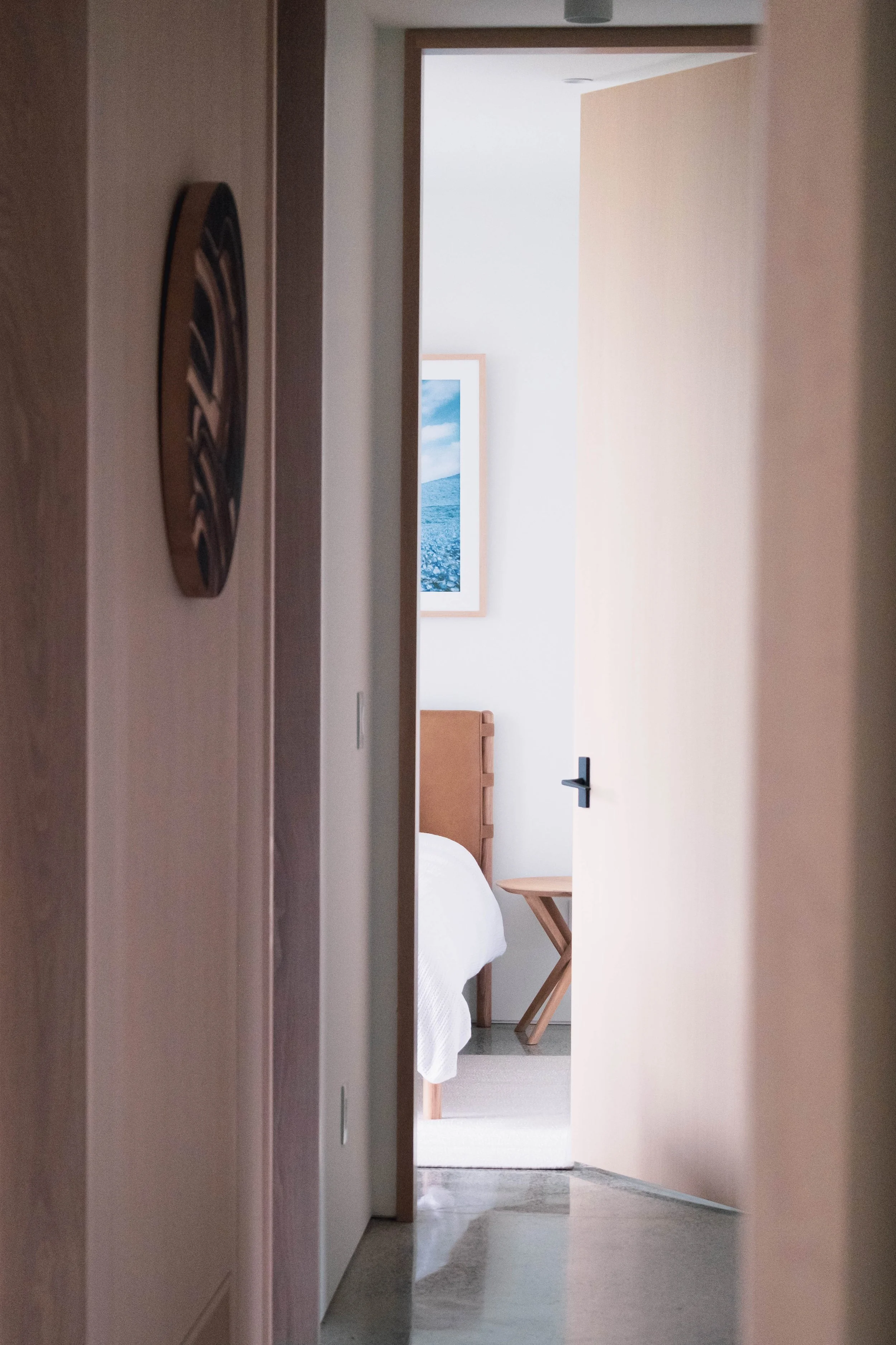 A view through an open door into a bedroom with a wooden headboard, a white bedspread, a wooden side table, and a framed ocean landscape on the wall.