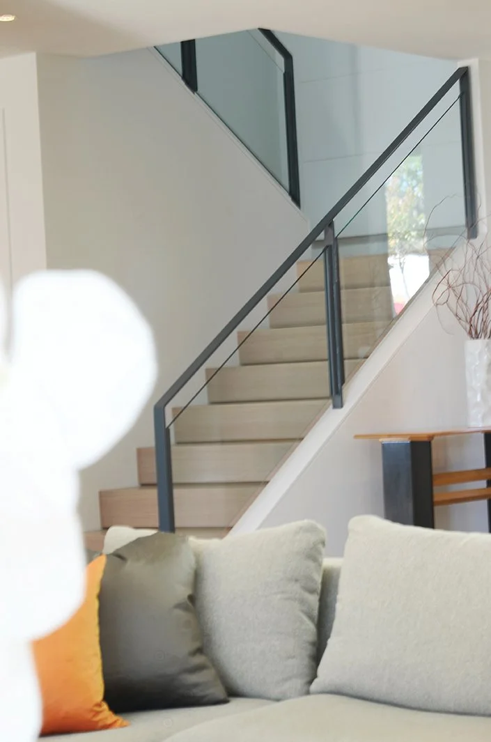 Interior of a modern house featuring a staircase with glass and metal railing, a beige sofa with colorful pillows, and a decorative vase with twigs on a wooden table.