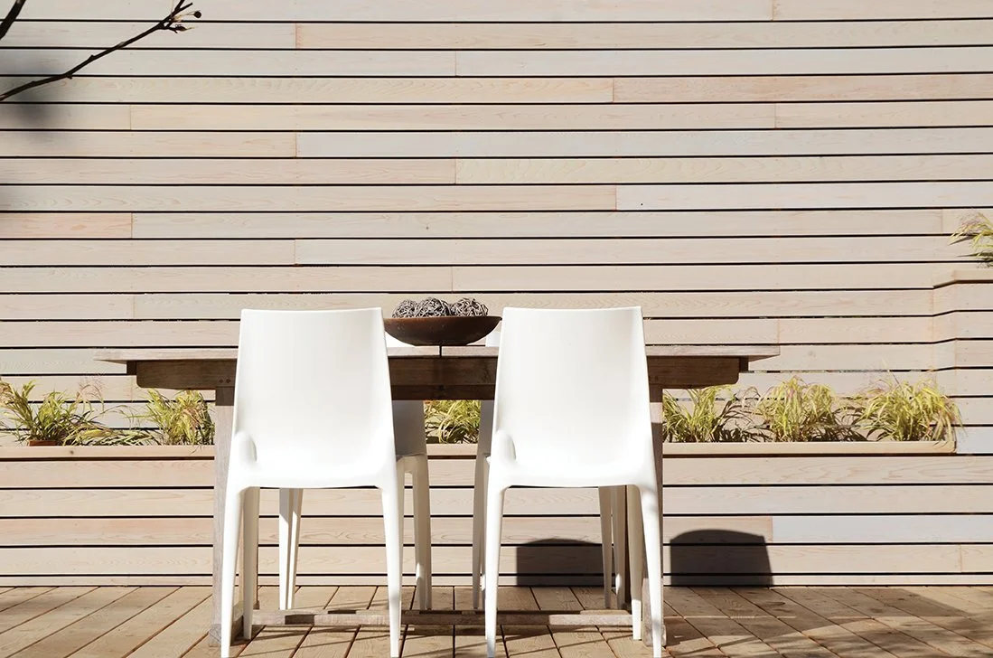 Outdoor patio with a wooden table, two white chairs, a bowl of decorative balls, and a wooden slat wall with a planled strip at the bottom.