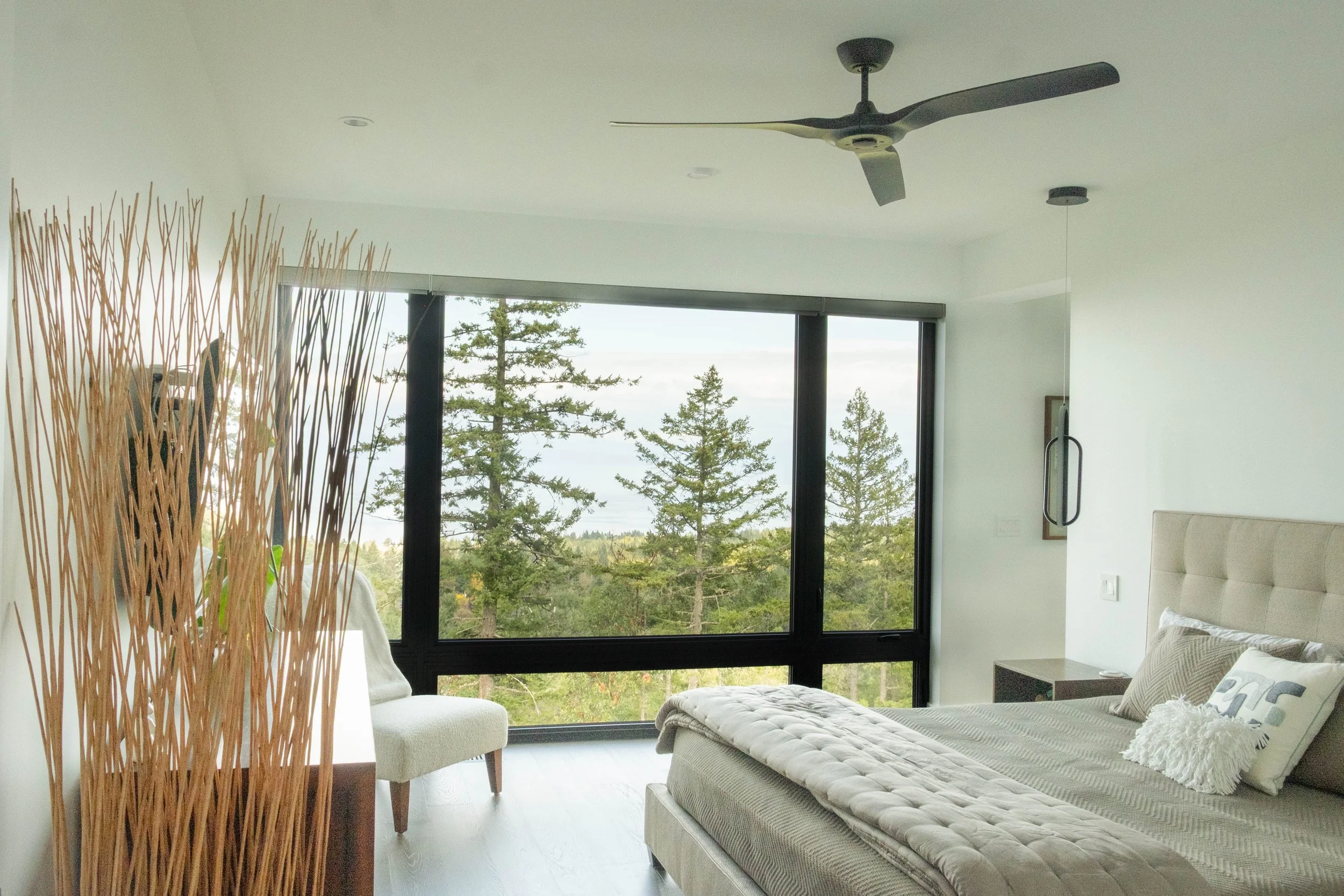 Modern bedroom with large window showing trees outside, bed with beige headboard and gray bedding, white chair, and ceiling fan.
