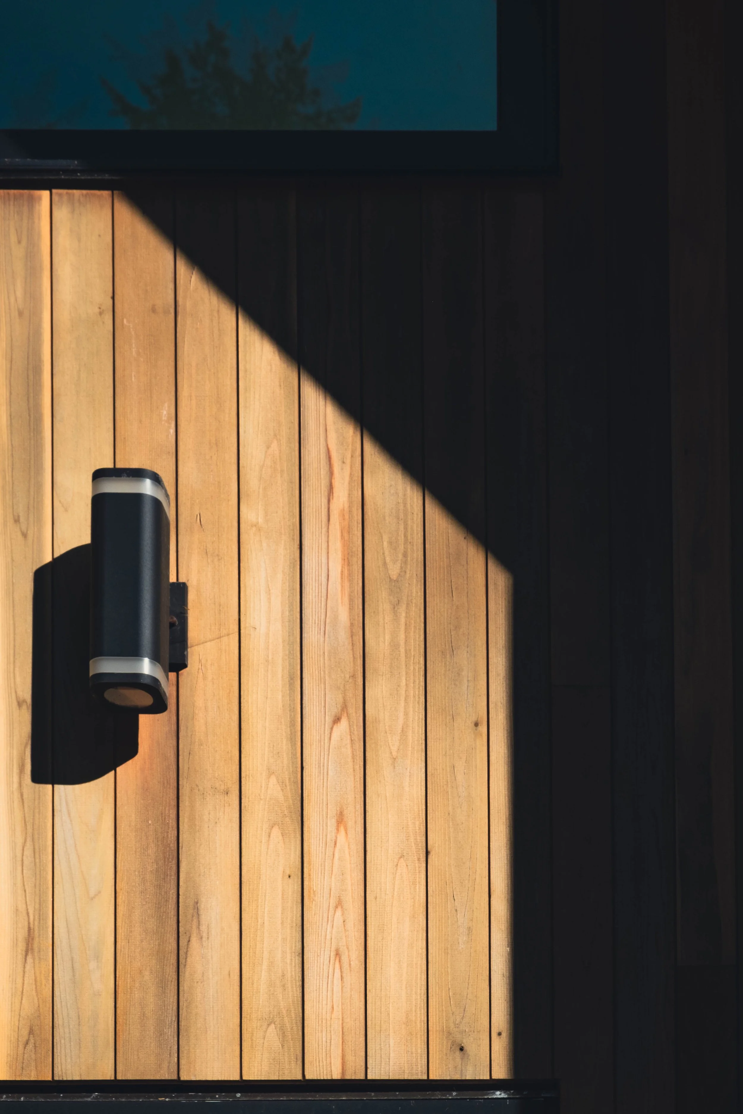 Wooden door with vertical planks, a modern handle, and a shadow cast across part of the door, with a view of the sky and trees visible in the upper window.