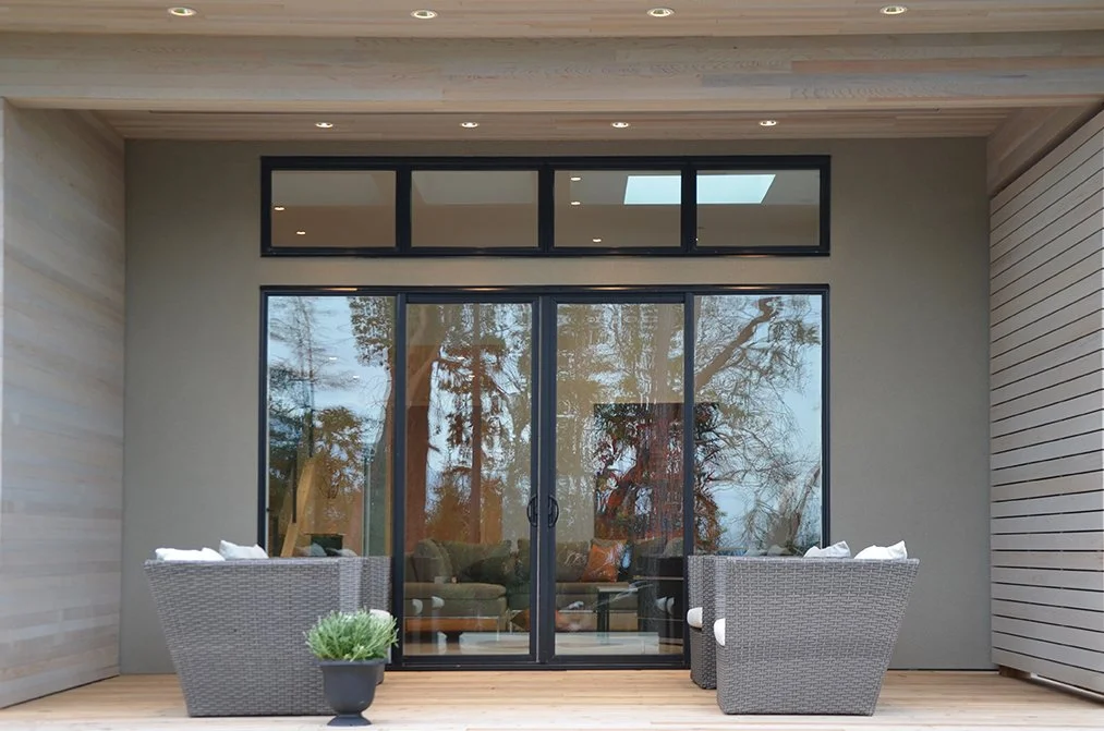 Modern house exterior with large glass sliding doors, outdoor seating with wicker chairs, a potted plant, and trees reflected in the glass.