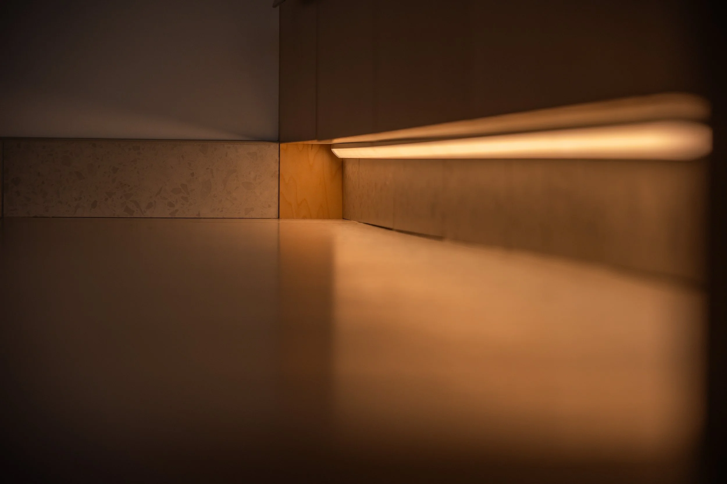 Close-up shot of a narrow space between a floor and a wall with warm lighting, showing the corner of a room or cabinet with wooden and stone surfaces.