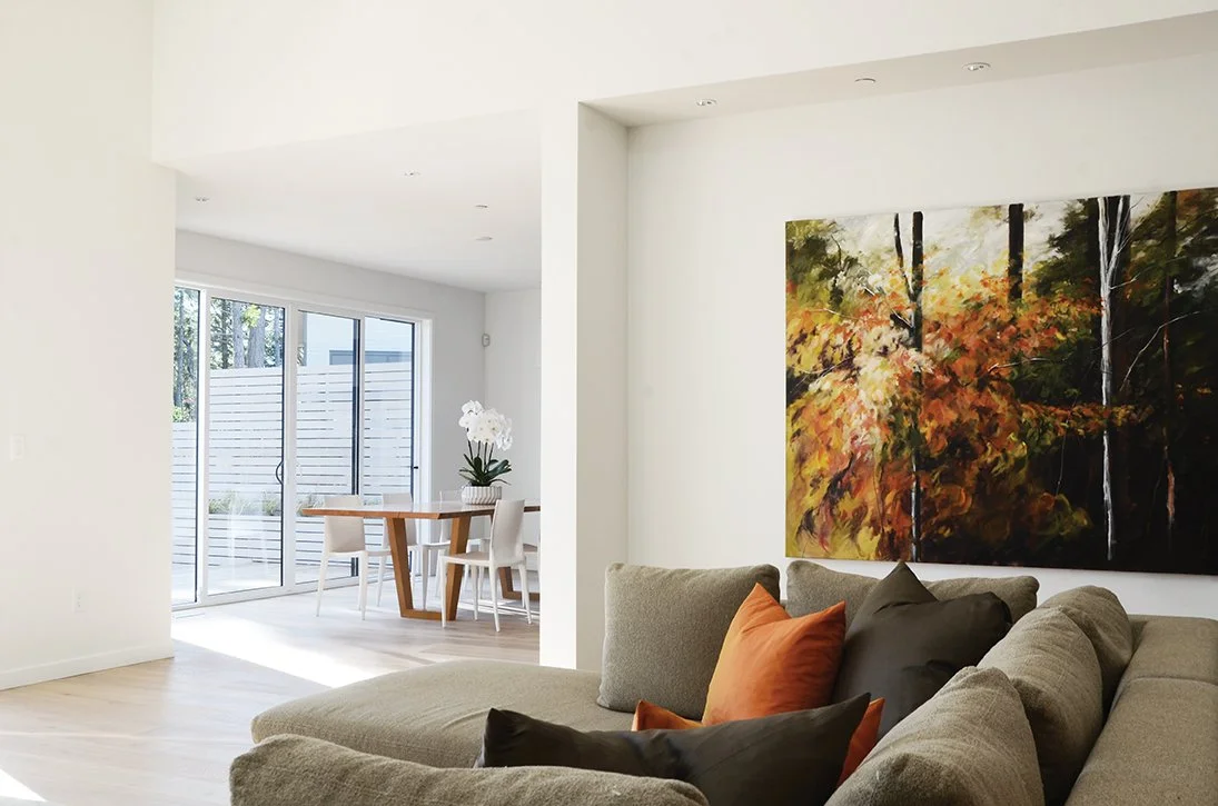 Living room with a beige sectional sofa and colorful pillows, a large landscape painting of trees with orange, yellow, and green leaves, and a dining area with a wooden table, white chairs, and a potted orchid near sliding glass doors leading outside