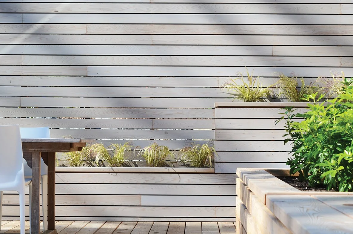 Outdoor patio with a wooden slat wall, planter box with tall grasses, and a potted green shrub.