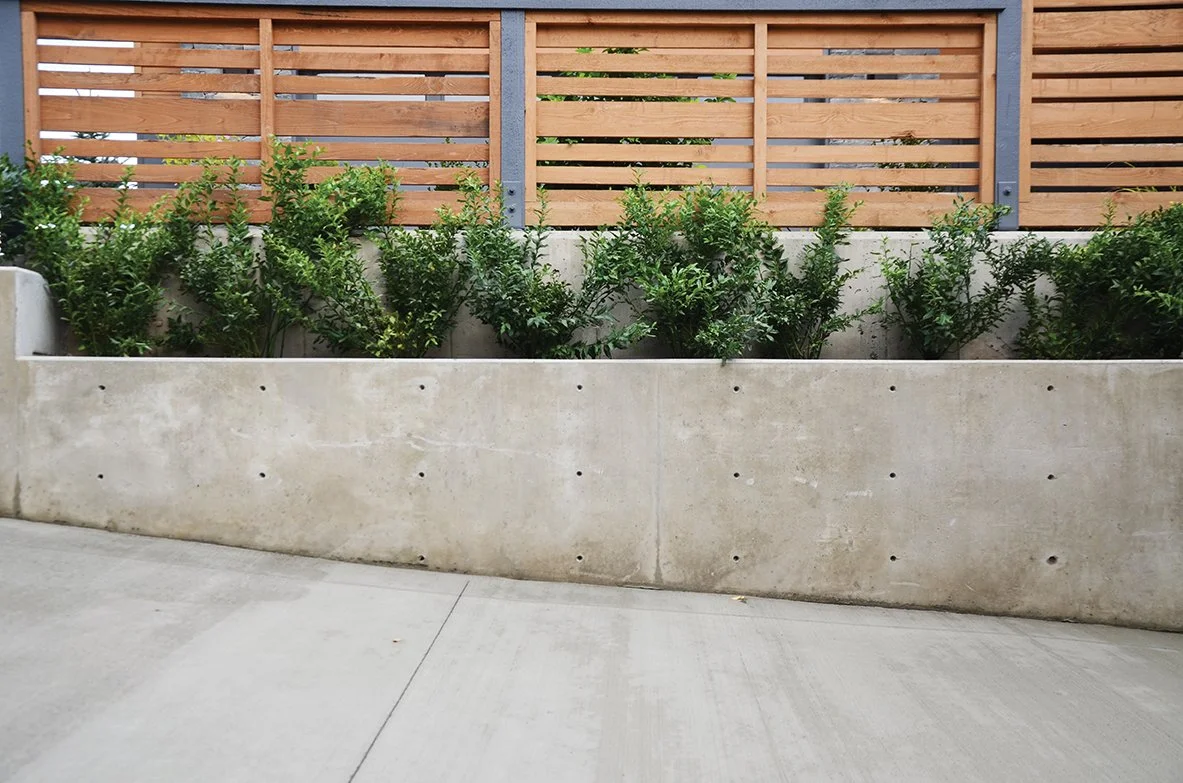 Concrete planter box with green shrubs and a wooden and metal fence backdrop in an outdoor setting.