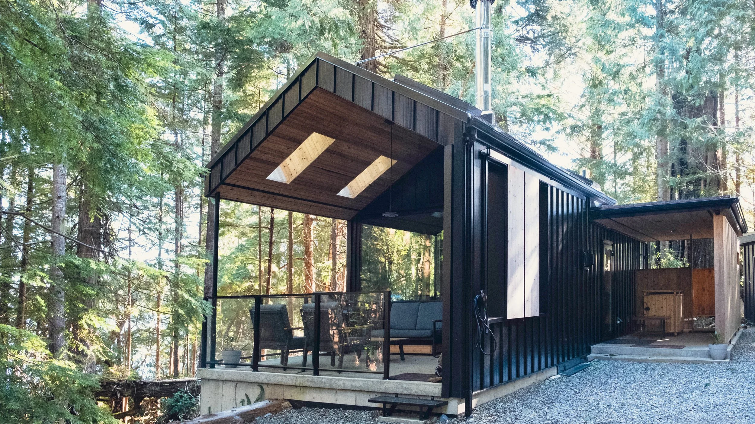 Modern black tiny house with a covered porch, sitting in a forested area with tall trees, featuring two skylights in the wooden ceiling of the porch.