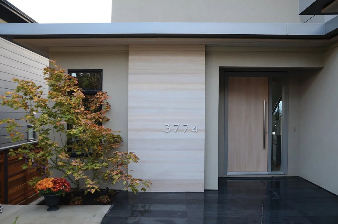 Exterior view of a modern house entrance with the house number 3774 on a light-colored wall, a wooden front door with a large handle, a small tree with green and orange leaves, potted plants, and a black tiled walkway.