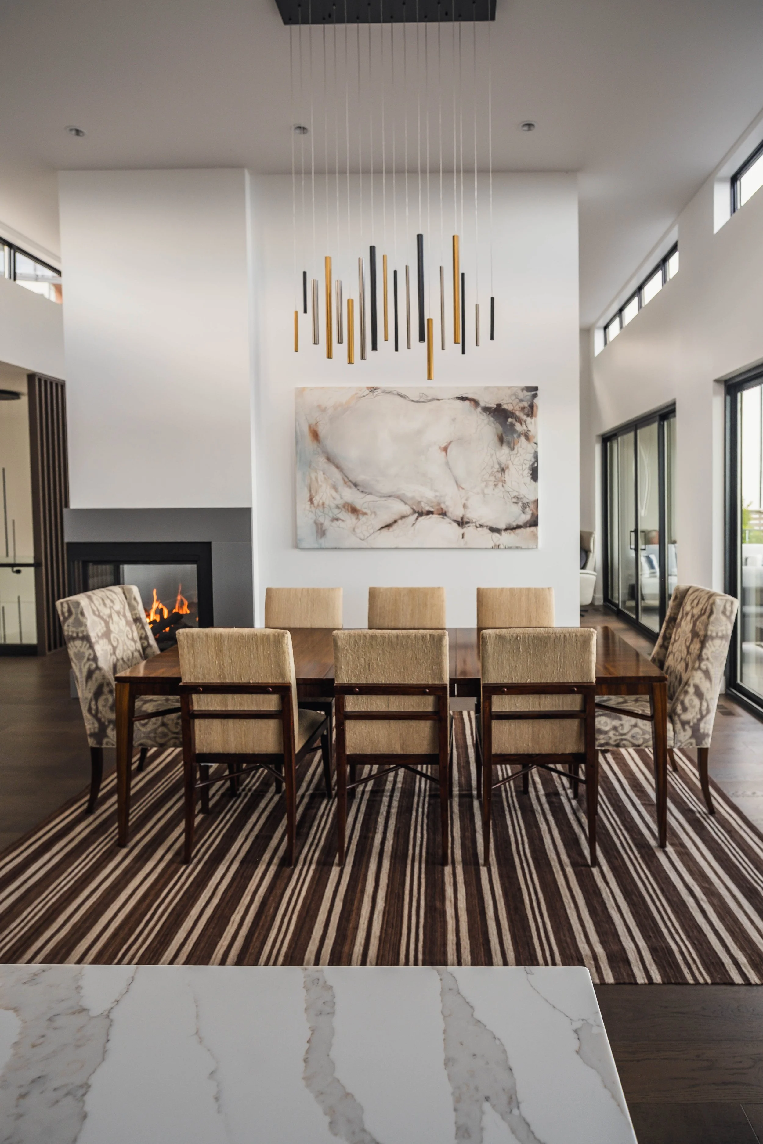 Modern dining room with a large wooden table, eight upholstered chairs, striped area rug, abstract painting on white wall, pendant light fixture, fireplace, and sliding glass doors leading to outdoor patio.