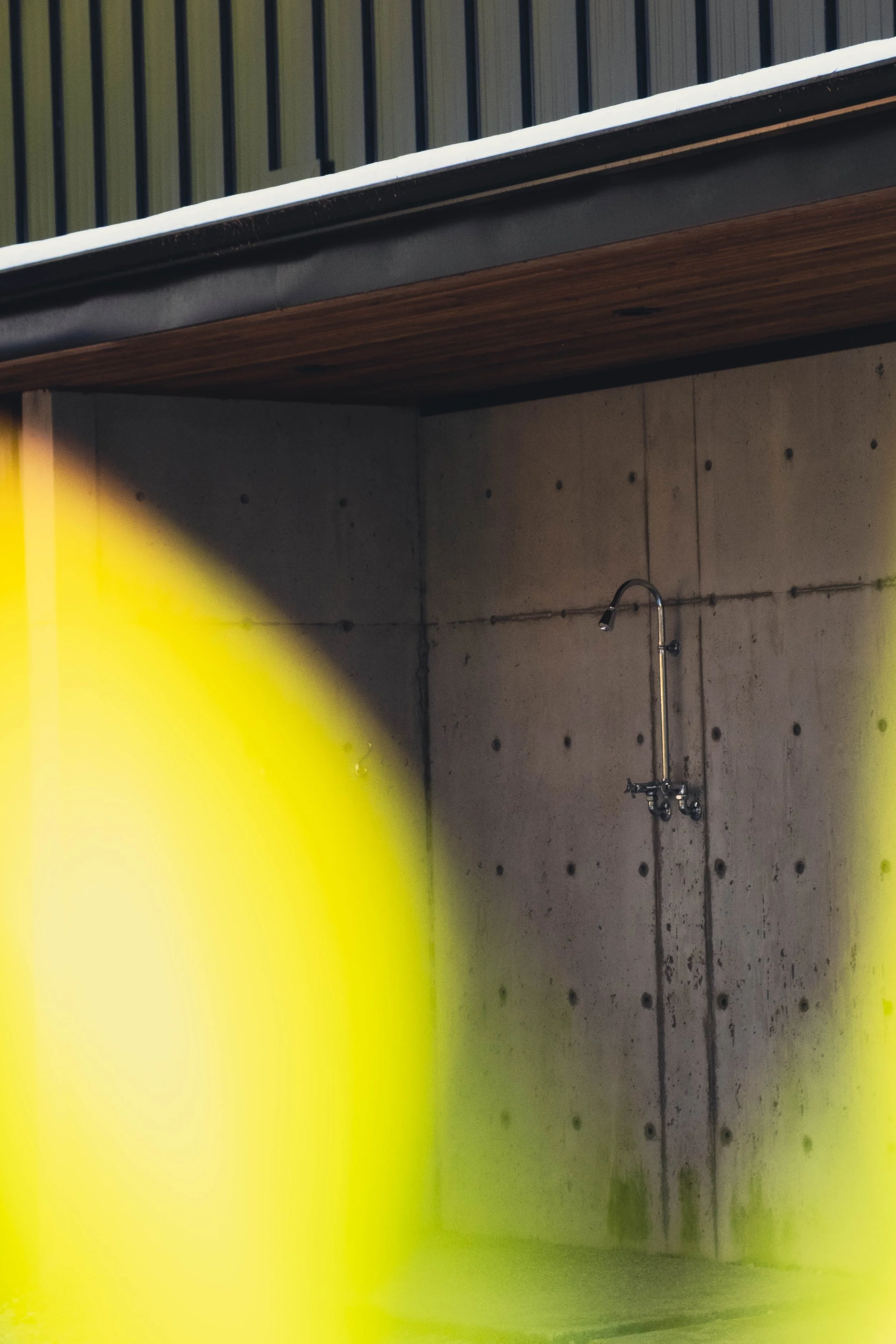 An outdoor concrete shower with a metal showerhead and faucet, partially obscured by a large yellow object or barrier in the foreground.
