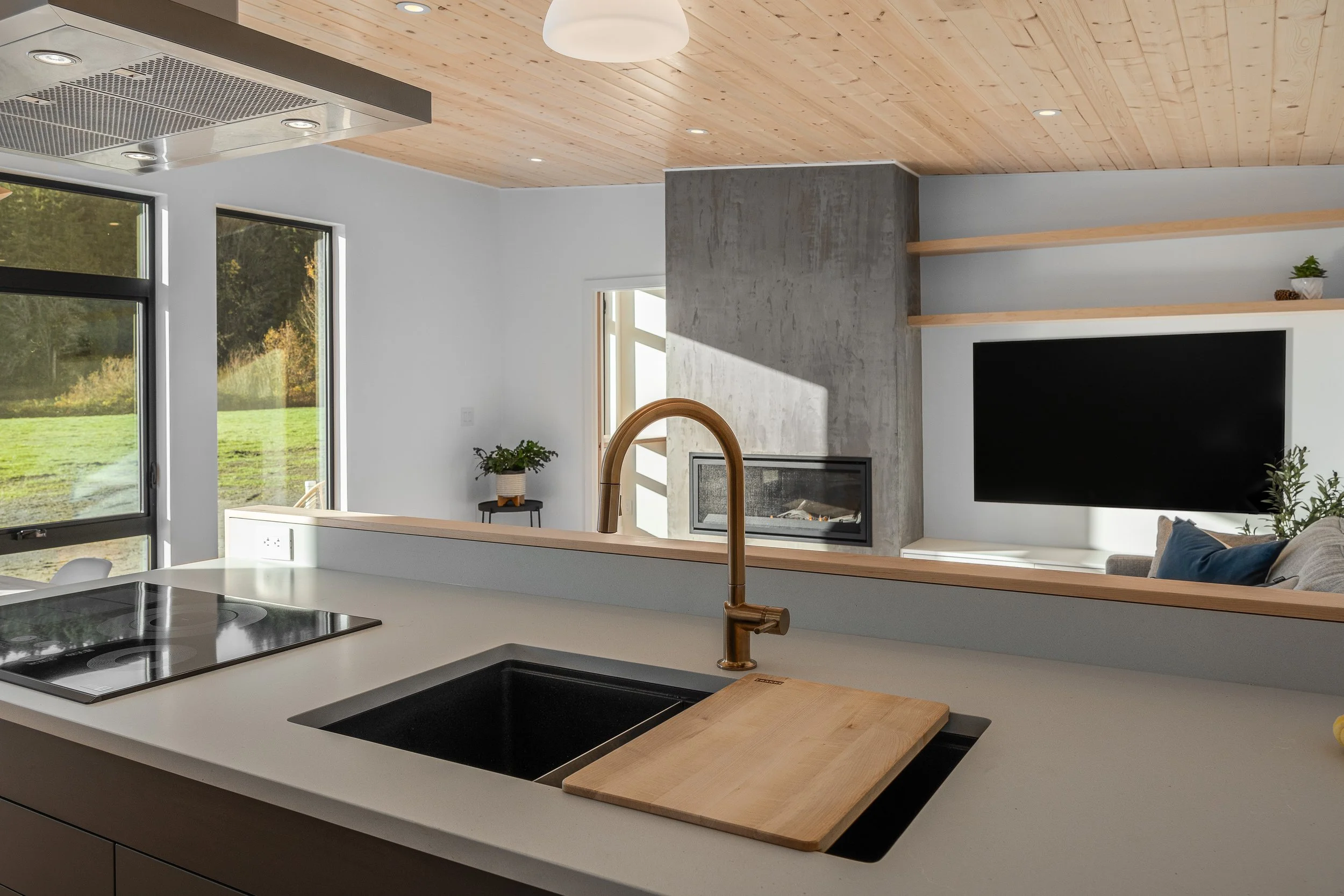 Modern open-concept kitchen with white countertop, black sink, and brass faucet. View into living room with fireplace, TV, and wooden shelves. Large windows showing outdoor green landscape.