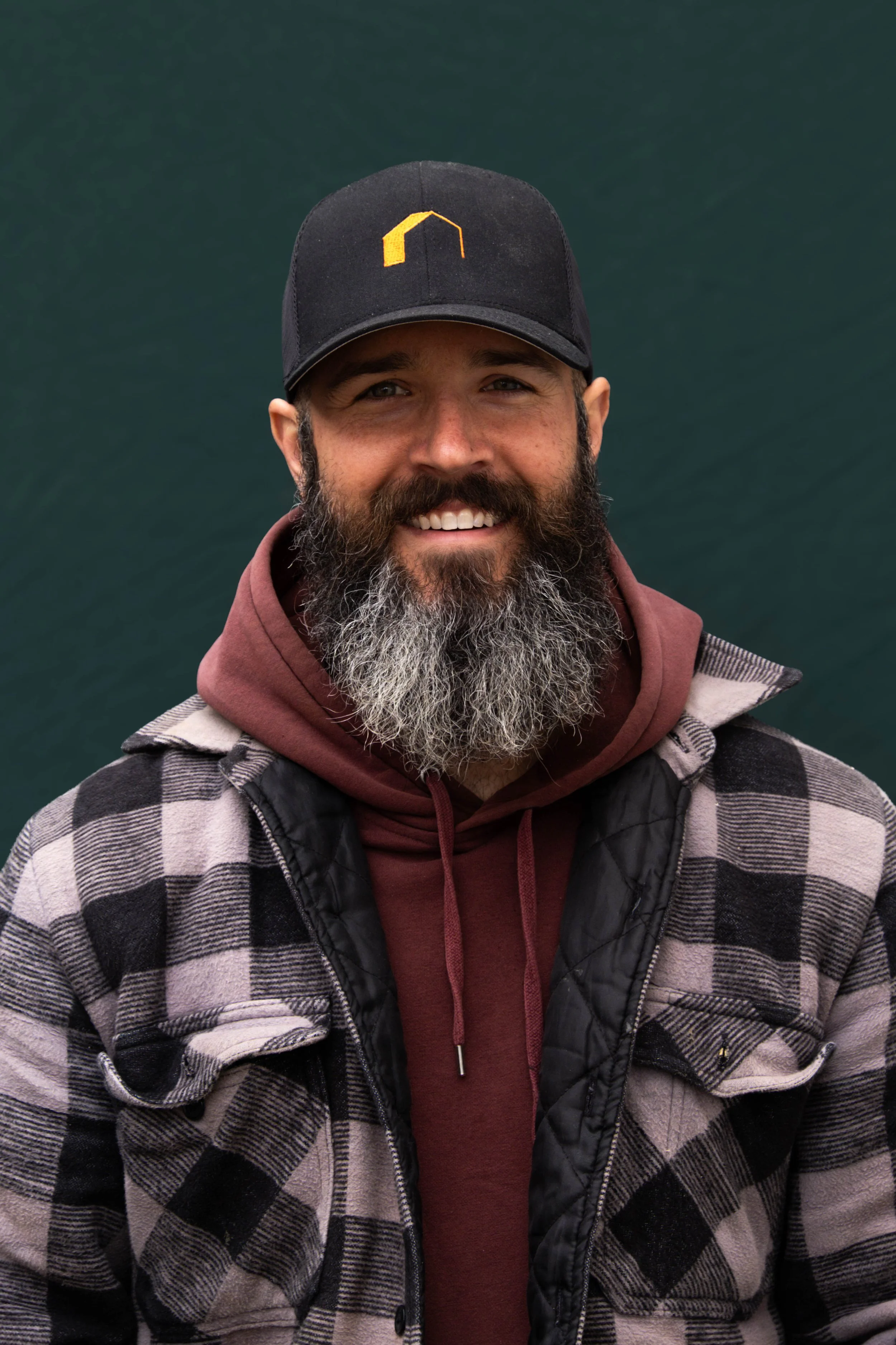 A smiling man with a beard and mustache, wearing a black cap with a yellow logo, a maroon hoodie, and a black and gray checkered jacket, standing outdoors with a dark green background.