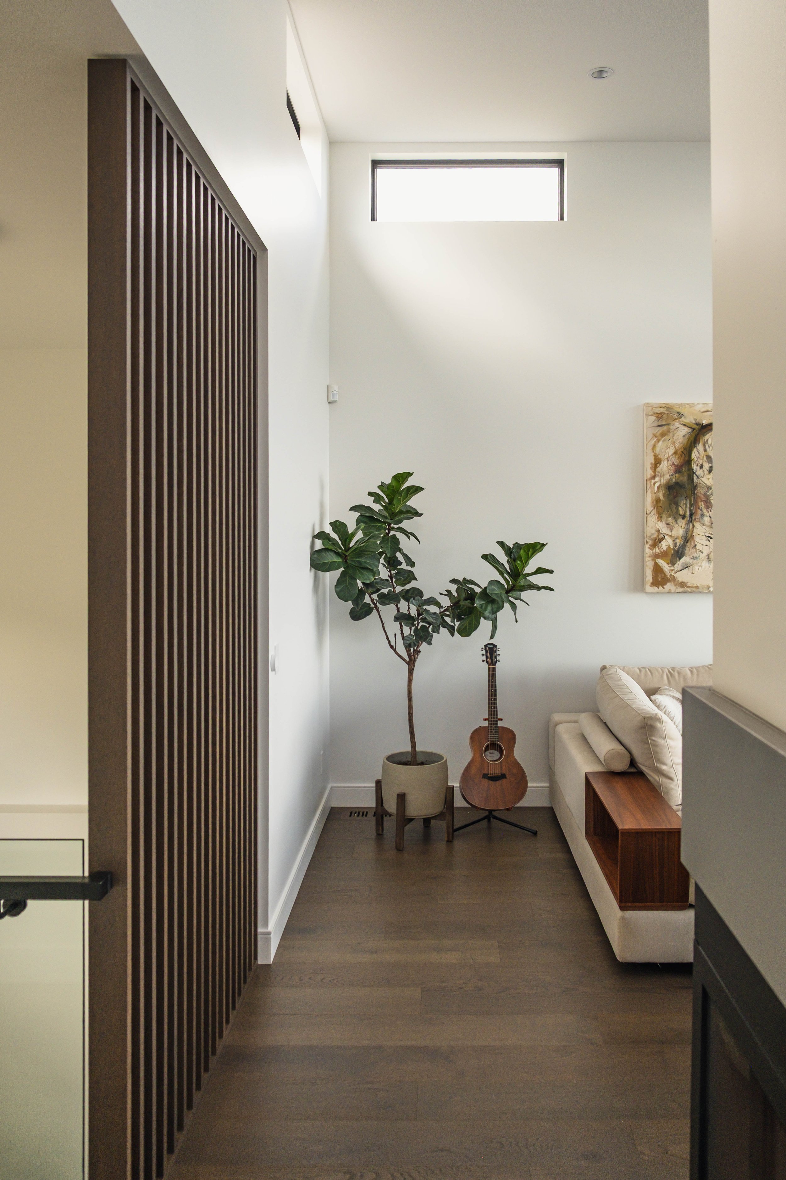A cozy living room corner with a tall potted plant, an acoustic guitar on stand, a white sofa, and a wooden side table, illuminated by natural light from a rectangular window high on the wall.