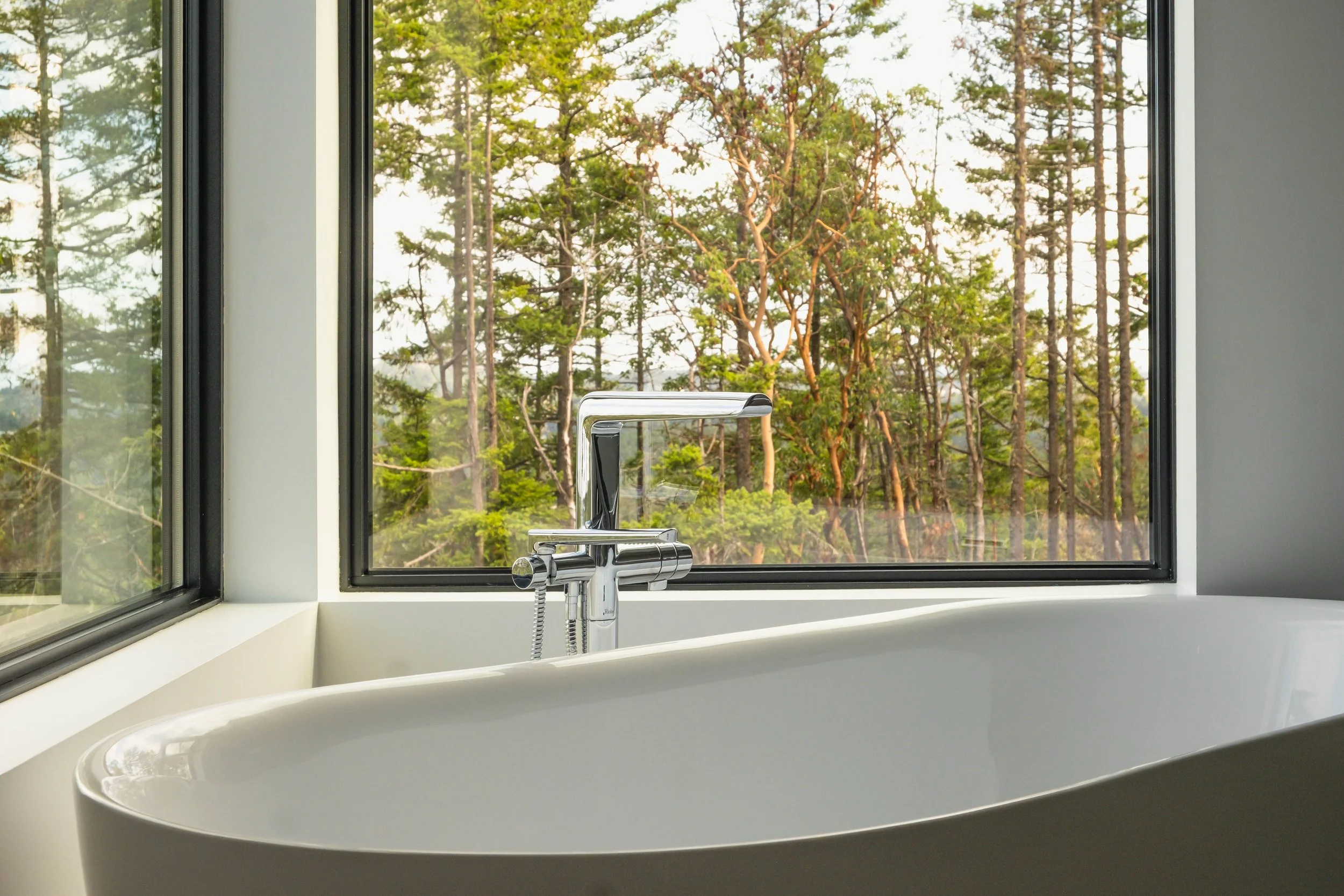 White bathtub next to a large window with a view of green trees and sunlight.