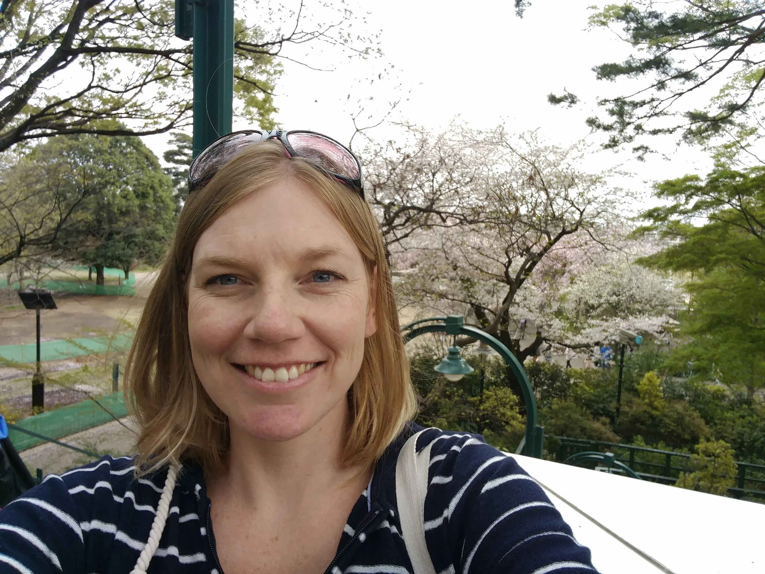 A woman smiling outdoors in a park with trees and blooming cherry blossoms in the background.