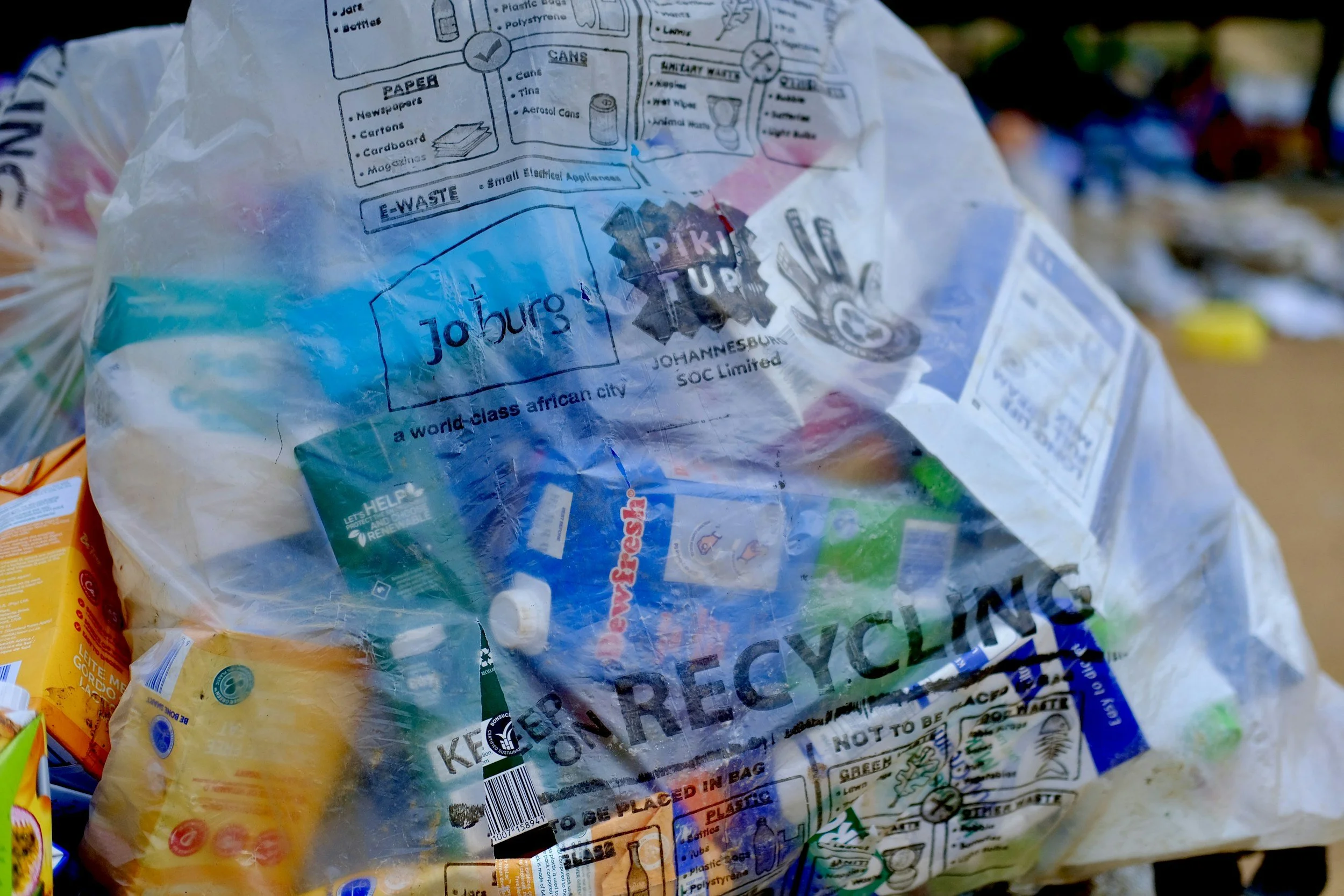 Close-up of a crumpled transparent plastic bag with recycling and disposal instructions printed in black, including mentions of paper, cans, glass, and plastic, and a barcode at the bottom, with blurry colorful items in the background.