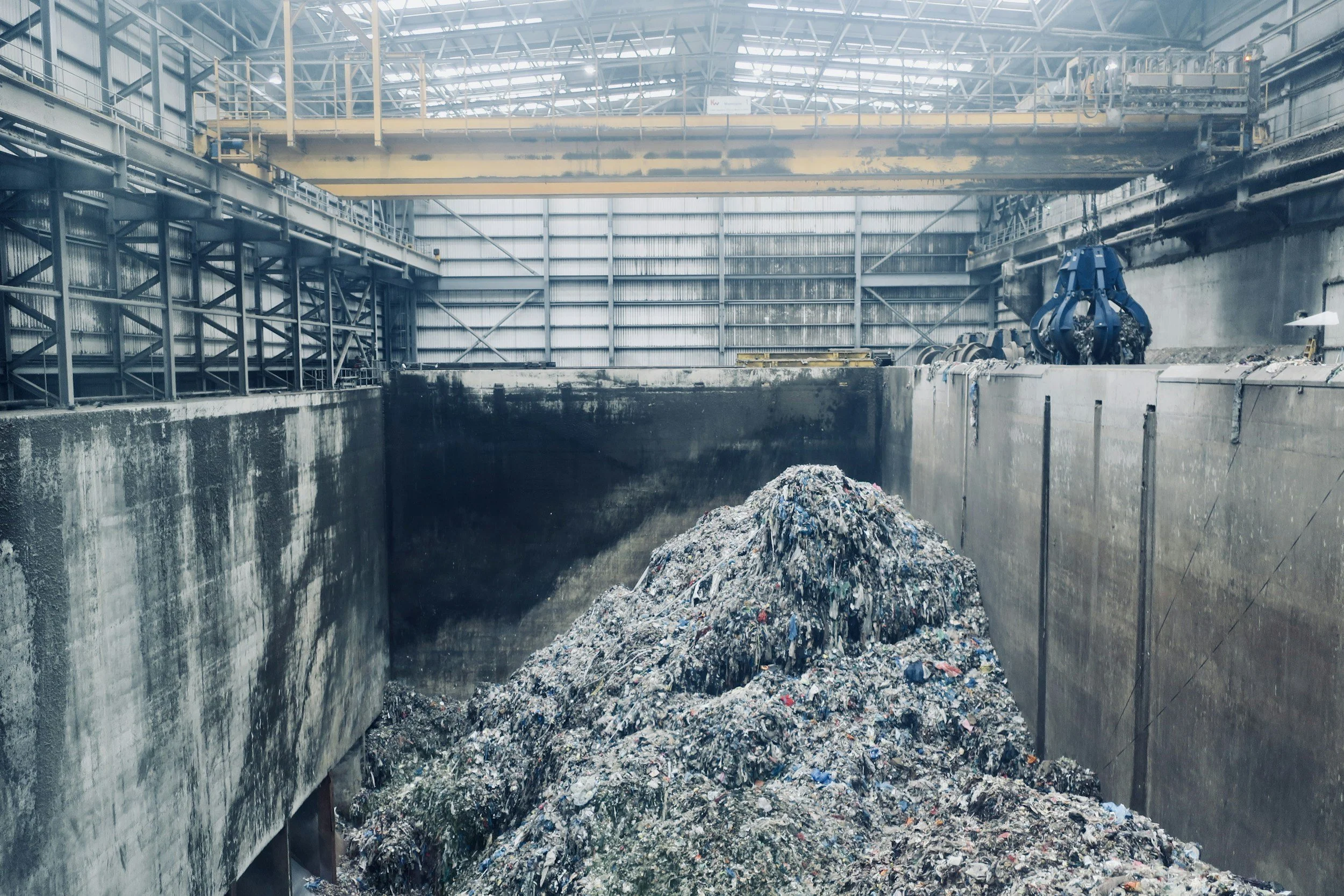 Inside an industrial waste processing facility with a large pit filled with compacted trash, surrounded by metal walls and overhead crane machinery.