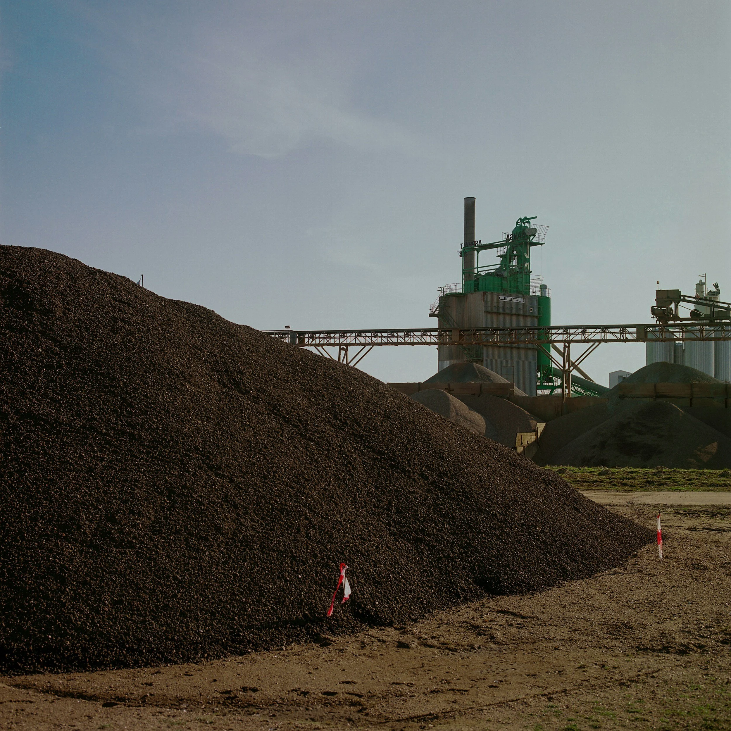 Industrial site with large piles of dark gravel or soil, factory structures including a tall chimney, and a conveyor system, under a clear blue sky.