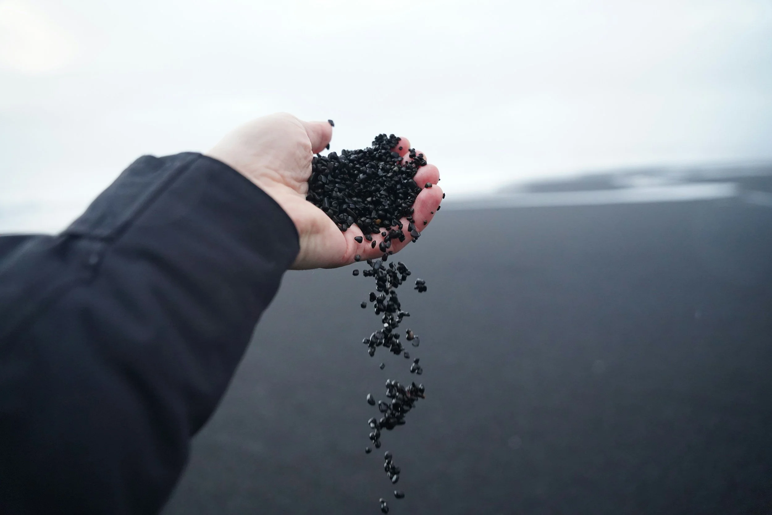 A hand holding small black stones or gravel with a cloudy sky in the background.