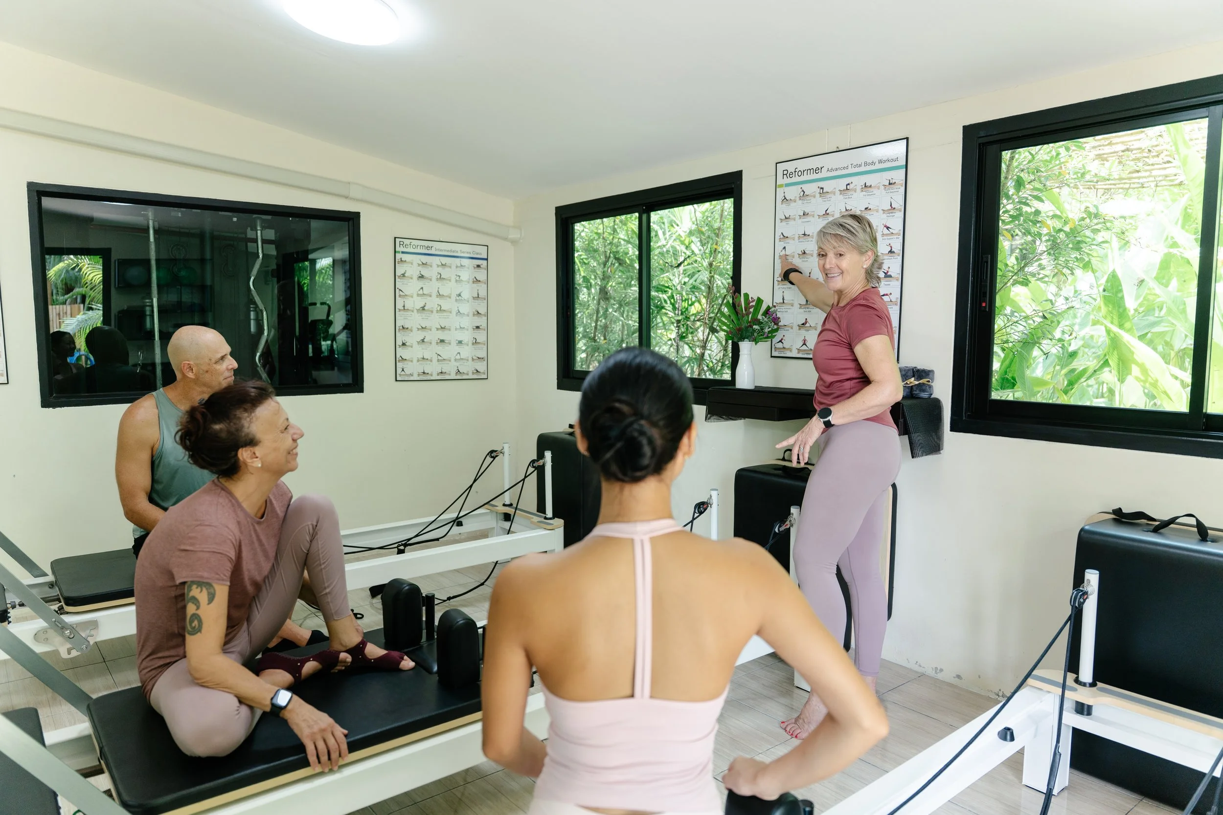 A fitness instructor leading a Pilates class in a studio with large windows and posters of Pilates exercises on the walls. The class participants are seated on specialized reformer machines, listening attentively.
