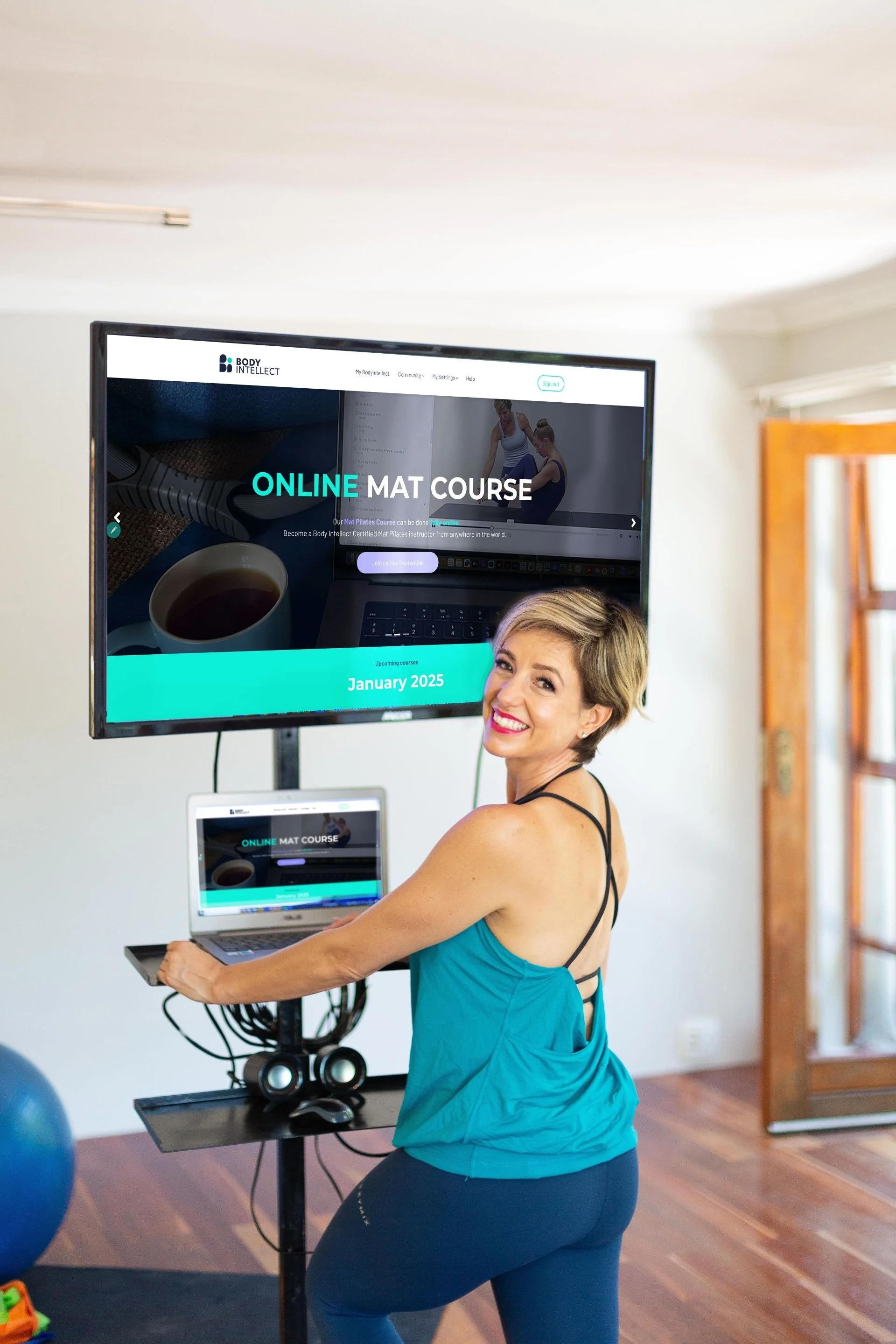 Theo Botha  holding a laptop in a home gym. She is standing next to a large monitor on a stand, which displays a website for an online mat course, with exercise images and text about the course scheduled for January 2025.