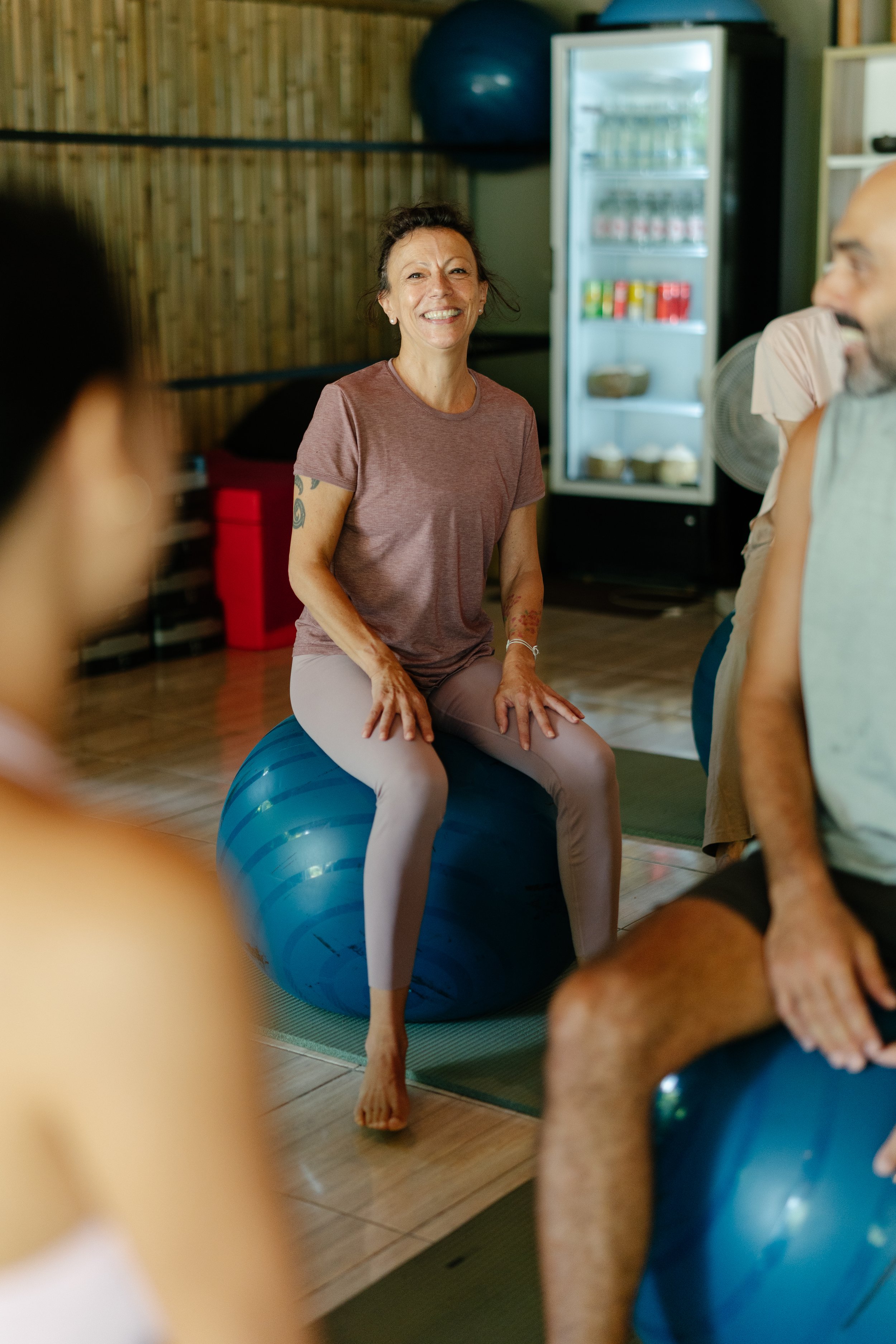 A woman smiling while sitting on an exercise ball during a fitness class surrounded by other participants in a gym or studio with wooden walls and a refrigerator in the background.