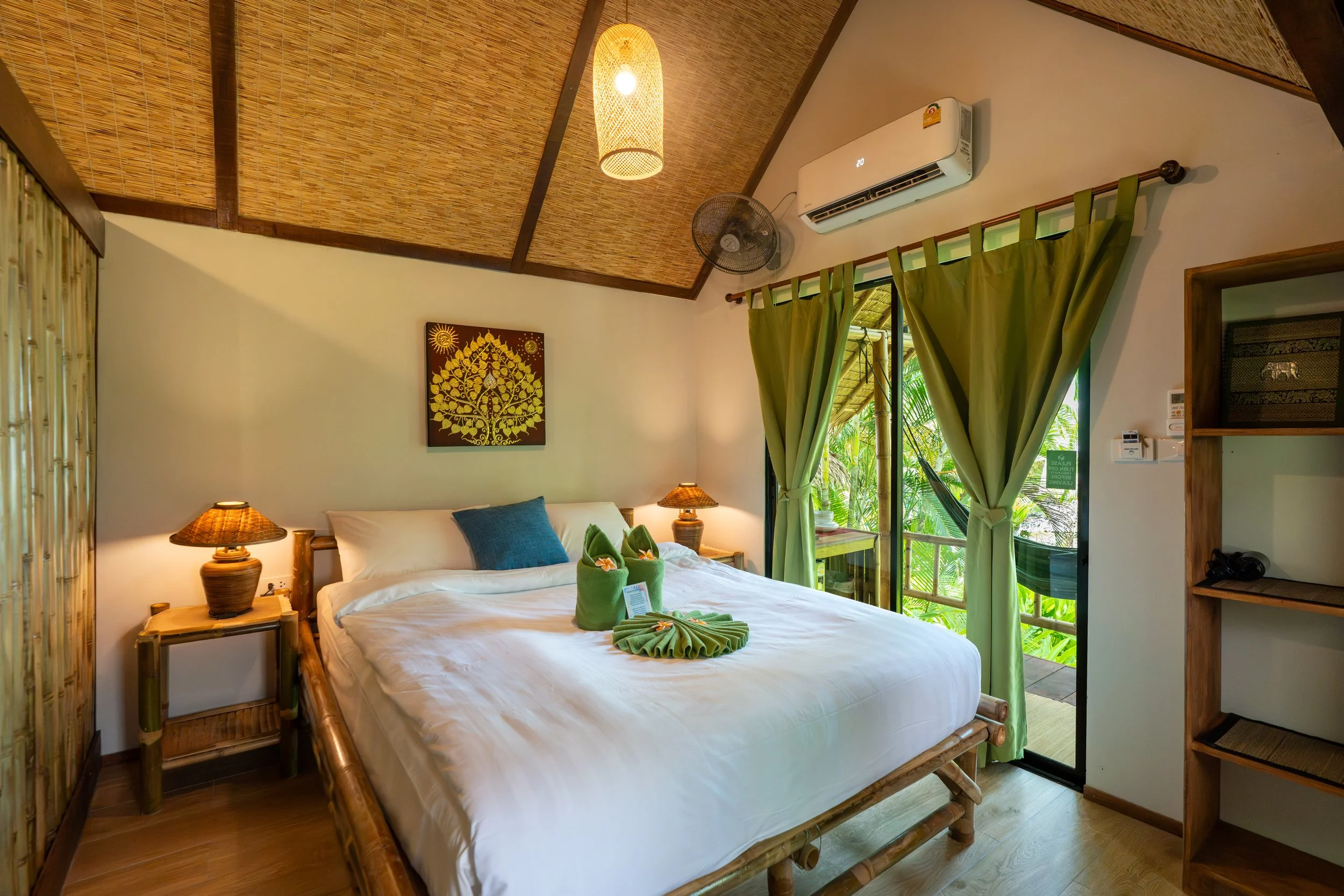 A cozy bedroom with a bamboo bed frame, white bedding, and a blue pillow. Two matching bedside lamps with woven shades sit on wooden nightstands. Green curtains frame a balcony door, revealing lush tropical foliage outside. The ceiling is thatched with bamboo, and a wall hanging with a gold tree design decorates the wall.