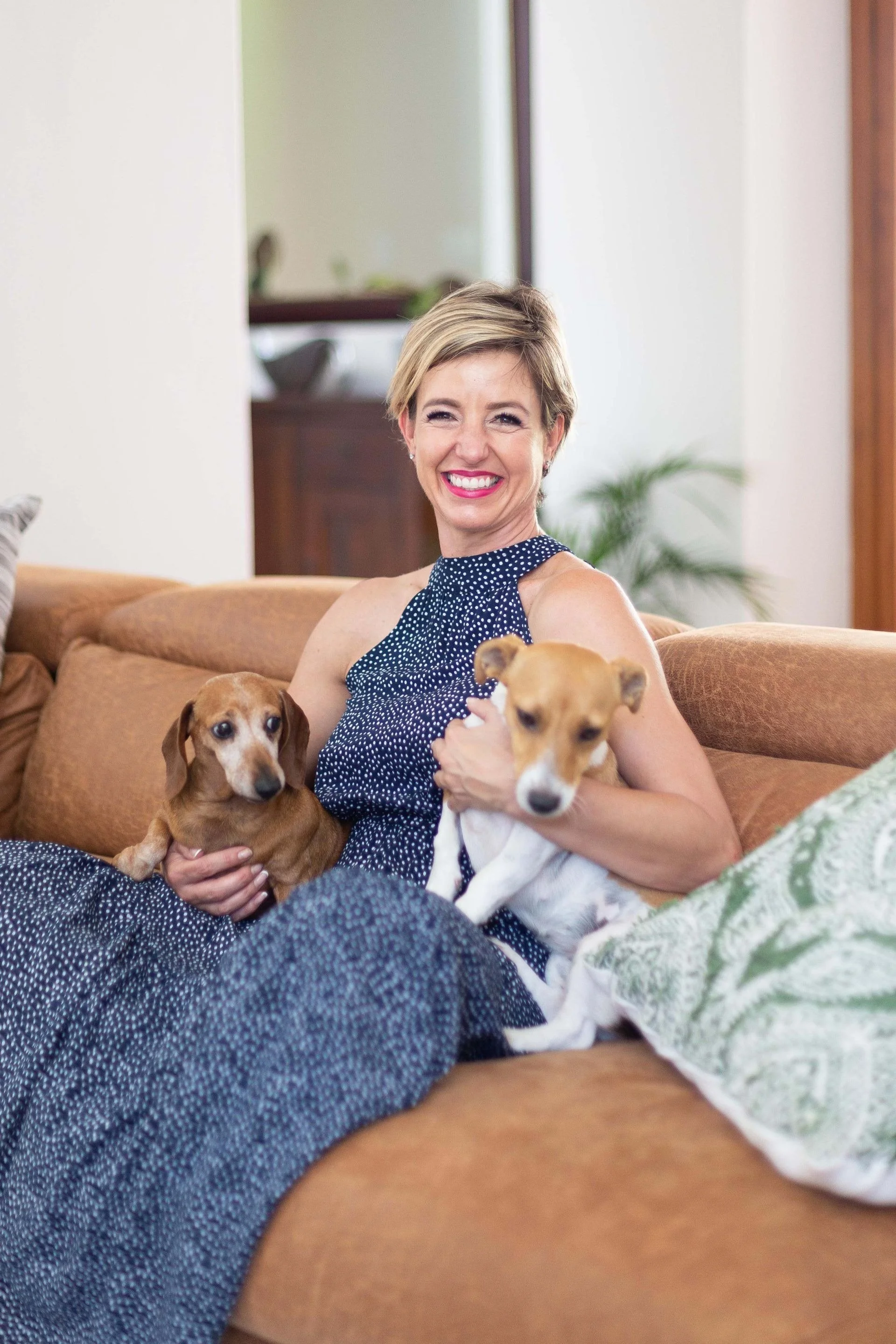 Theo Botha sitting on a tan couch, smiling while holding two small dogs, a brown one and a white one with patches, in a cozy living room with a mirror, plants, and a wooden cabinet in the background.