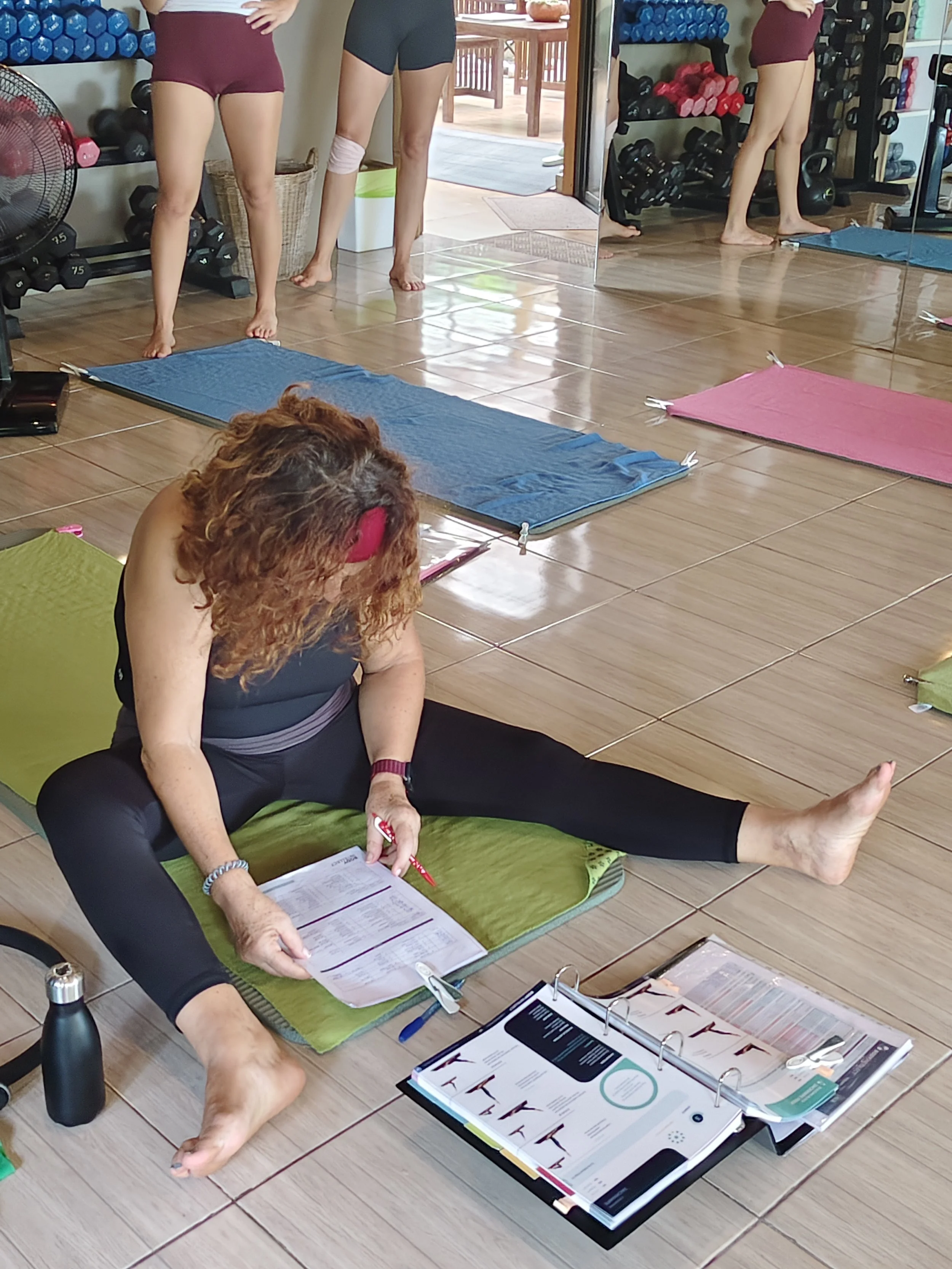 A group of women participating in a yoga or fitness class in a studio, with exercise mats on the floor, some standing and one sitting on a green mat with headphones and reading material.