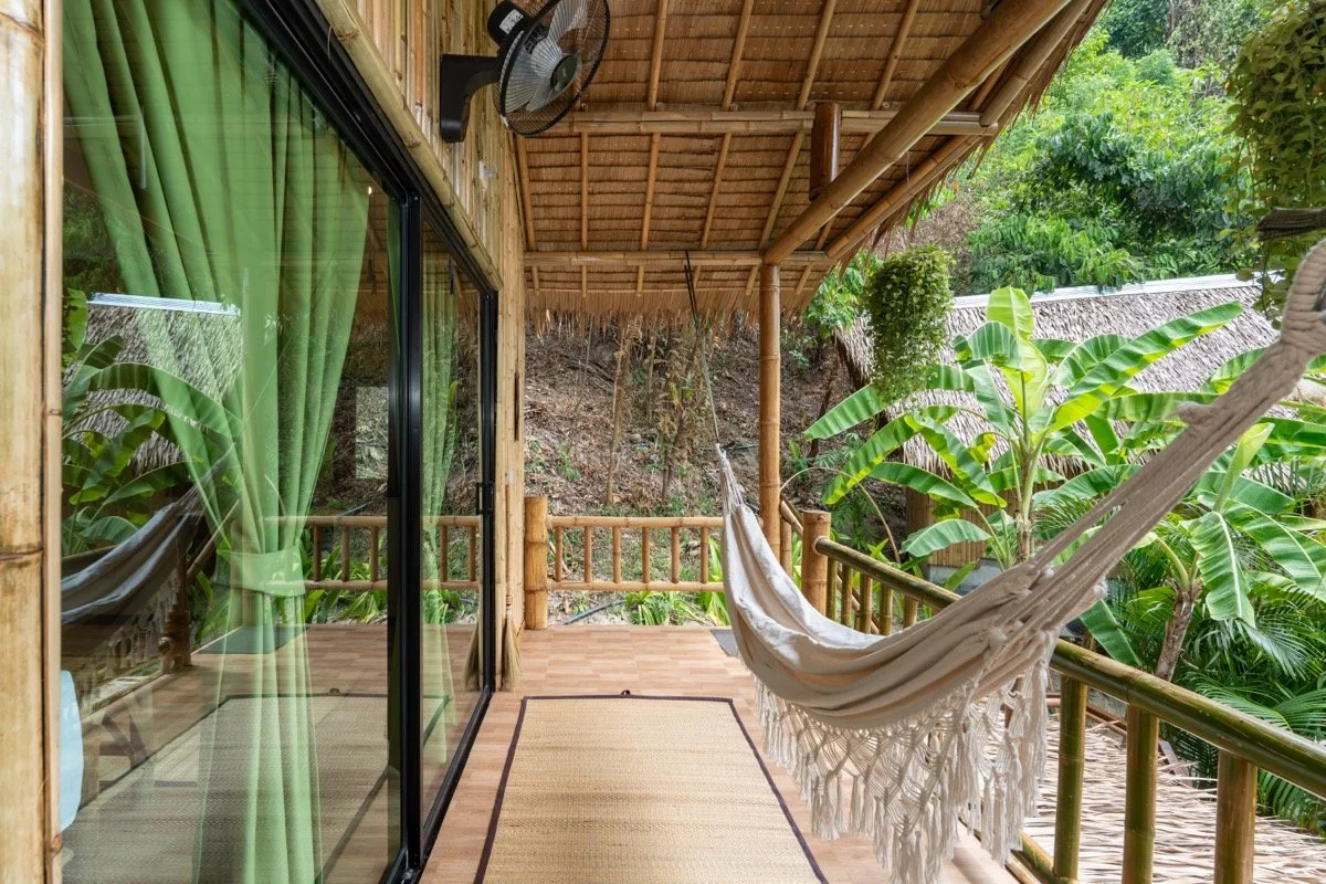 View of a wooden porch with green curtains, bamboo railing, hanging hammock, ceiling fan, and lush green tropical plants outside.