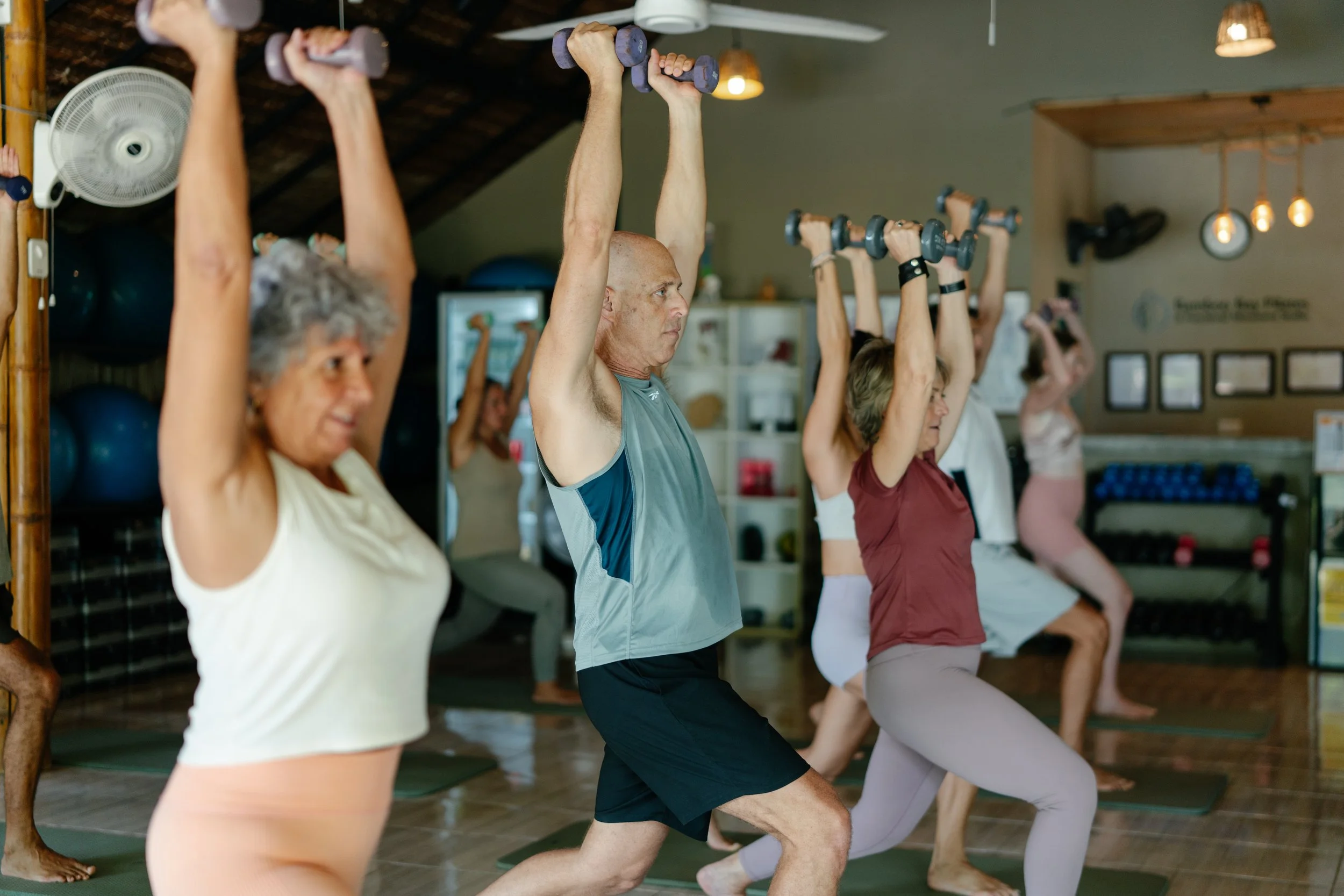 Group of diverse seniors participating in a pilates or exercise class, holding dumbbells overhead in a studio with wooden walls and warm lighting.