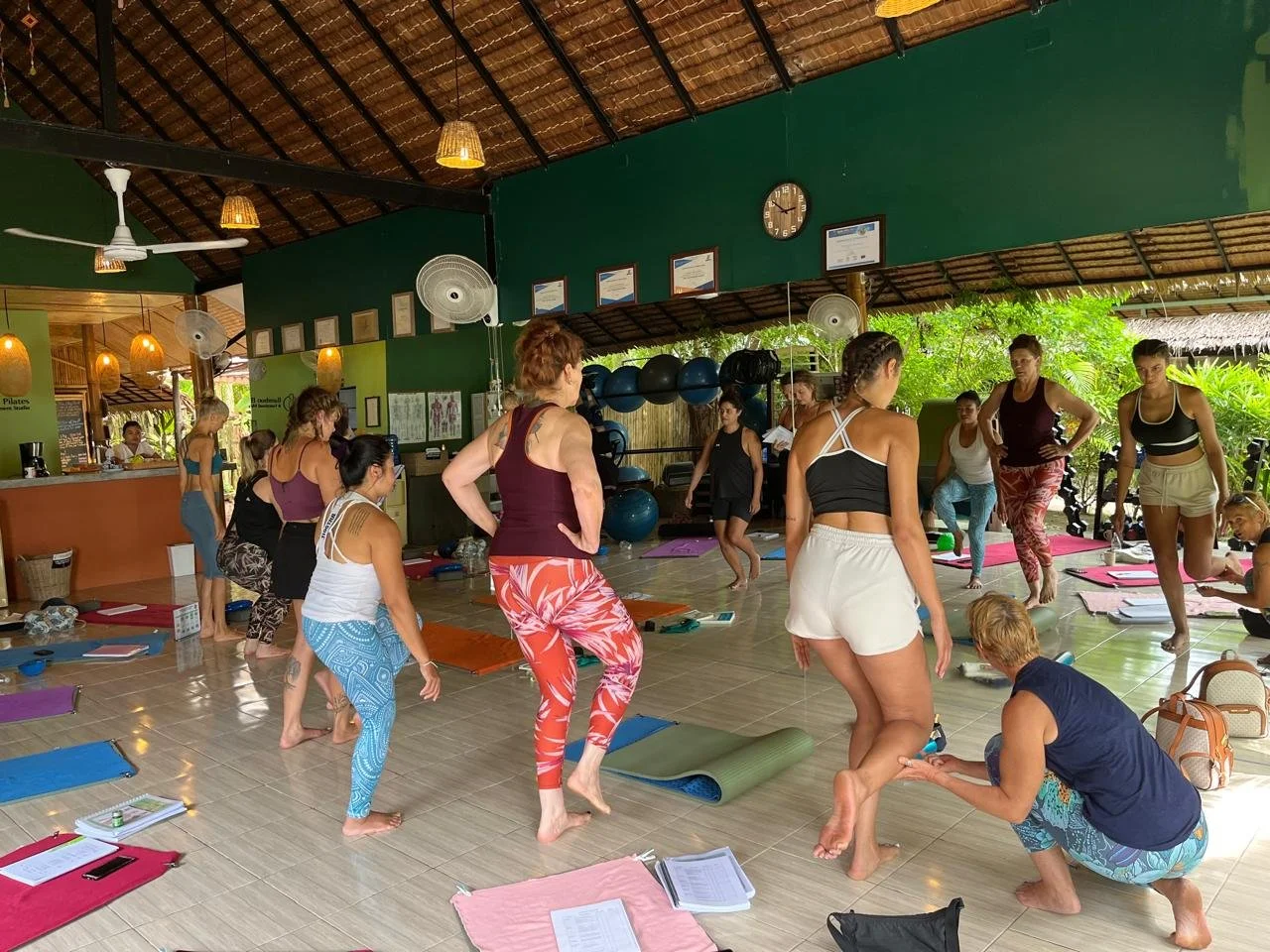 People participating in a yoga class in a tropical, open-air studio with green walls, mats, and hanging lamps.