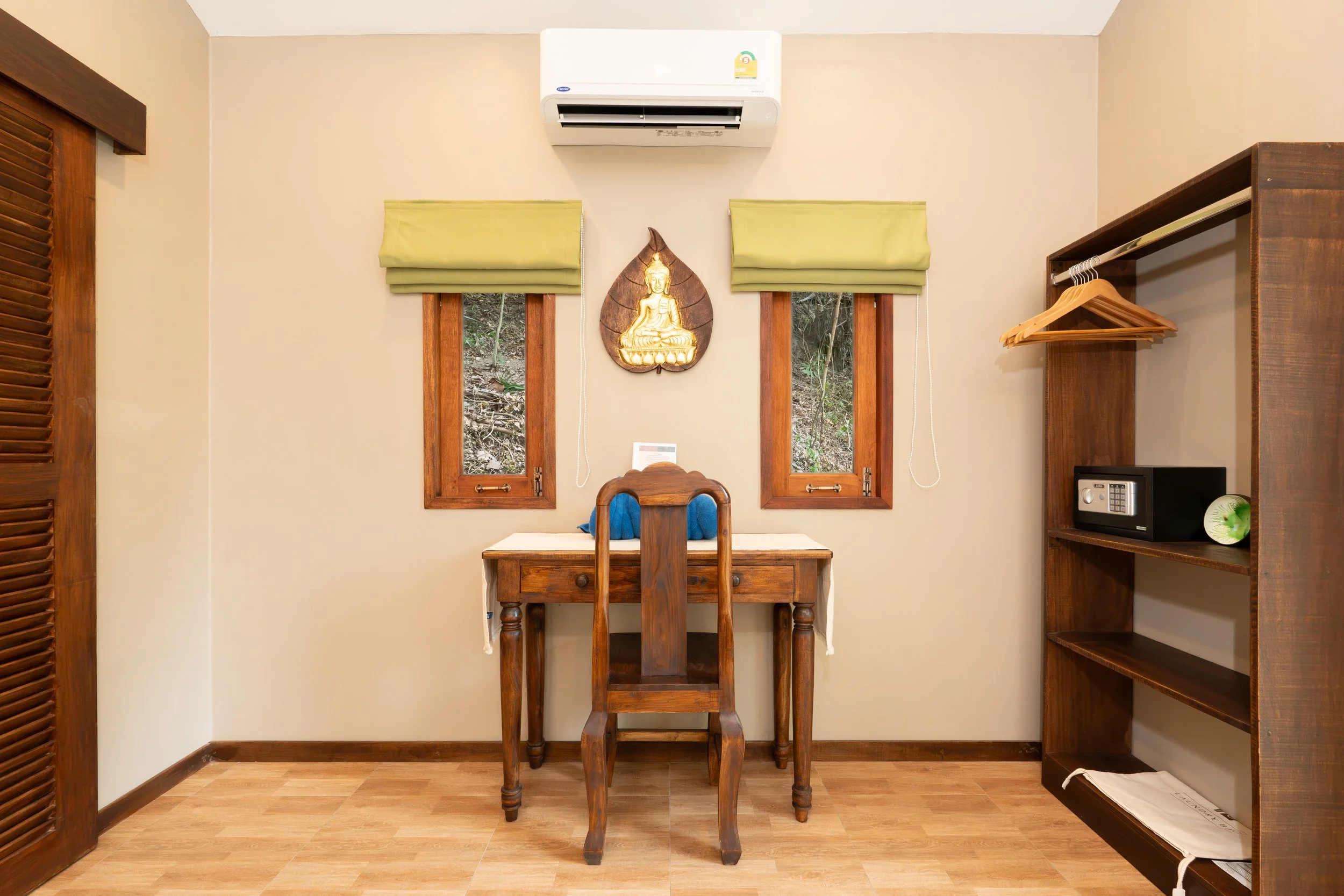 A hotel room with two small windows, wooden furniture, and a decorative Buddhist wall hanging featuring a golden Buddha figure. There is a small desk with a chair in front of the windows, a bookshelf on the right, and an air conditioning unit above.