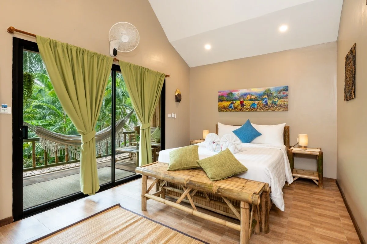 A bedroom with a bed decorated with white sheets, a blue pillow, and two green pillows. There is a rustic wooden bench at the foot of the bed. Two bedside tables with lamps are on either side. The room has beige walls, a wall art above the bed, and a sliding glass door with light green curtains leading to a balcony with a hammock and lush green tropical plants outside.