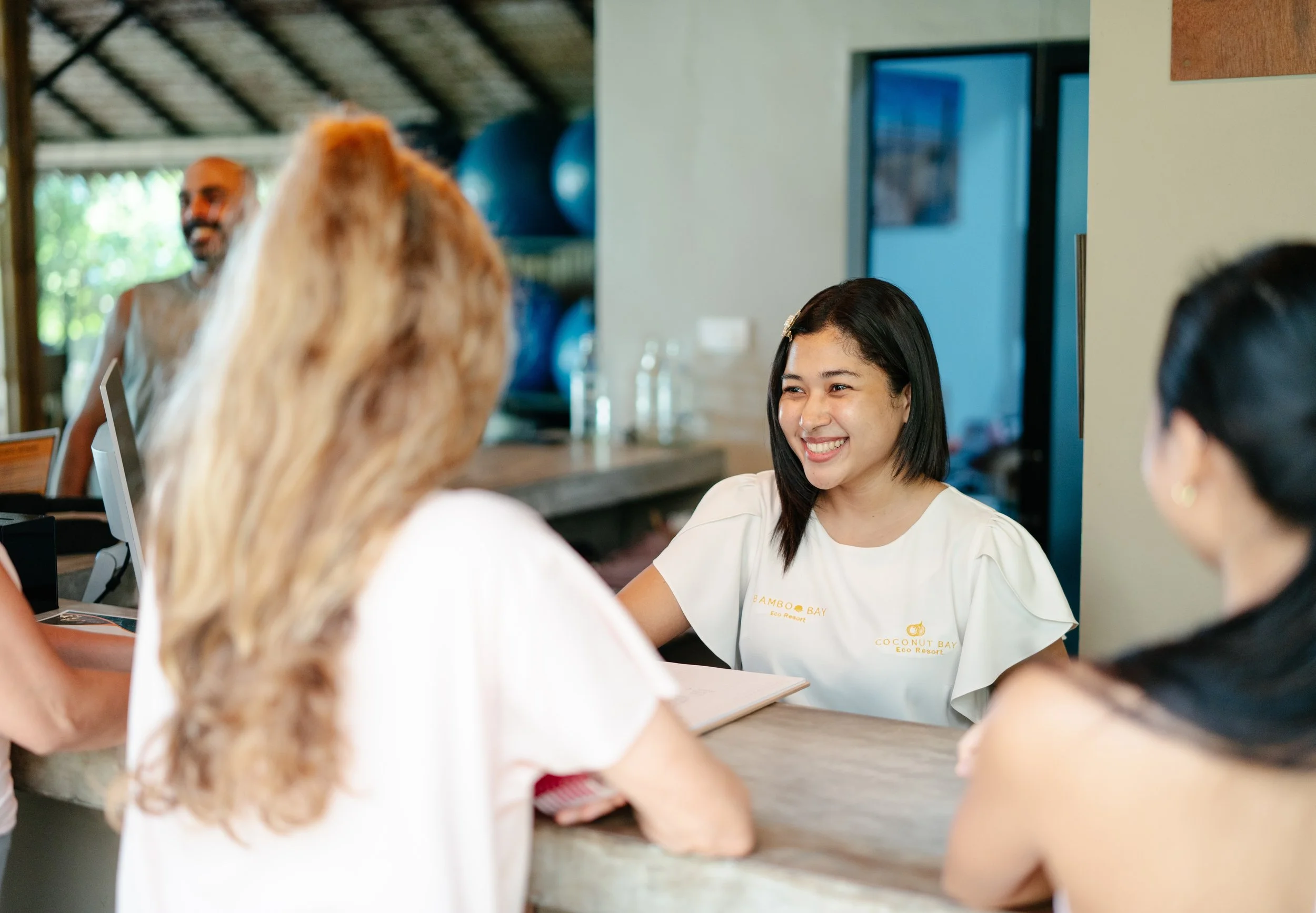 A group of people sitting around a wooden table, engaging in a conversation, with a young woman smiling, wearing a white shirt with 'BAMBOO BAY Eco Resort' written on it.