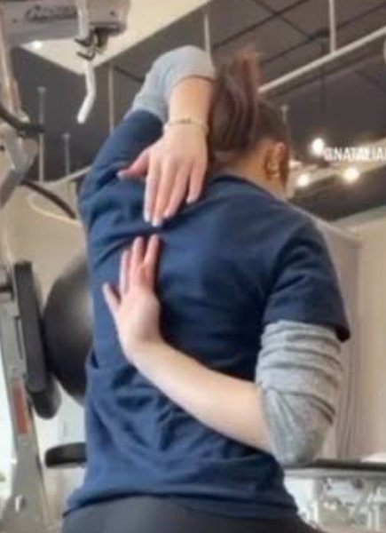 A woman exercising in a gym, stretching with her body bent forward and one arm over her head.