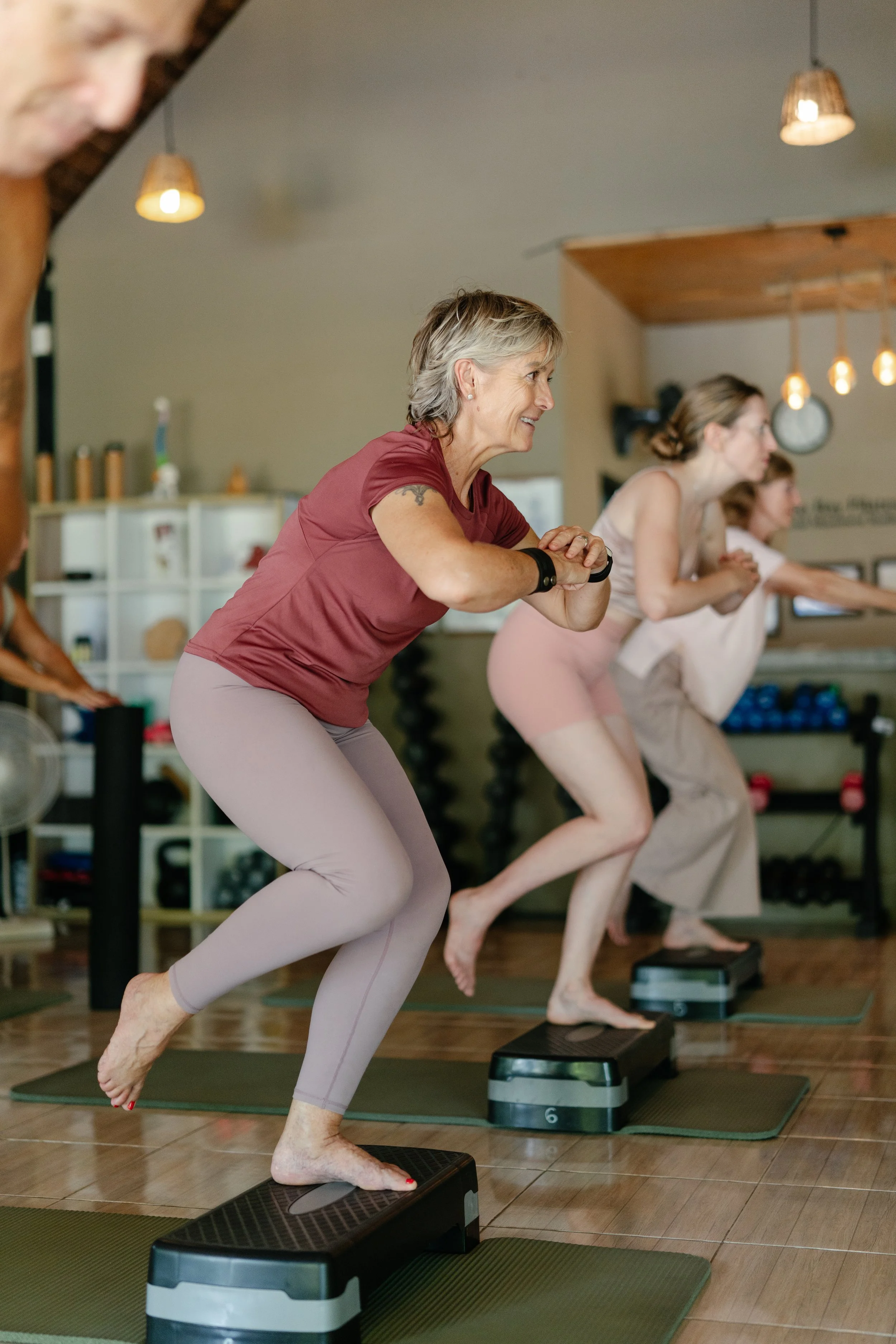 Women participating in a step aerobics class, standing on step platforms with their hands clasped and smiling.