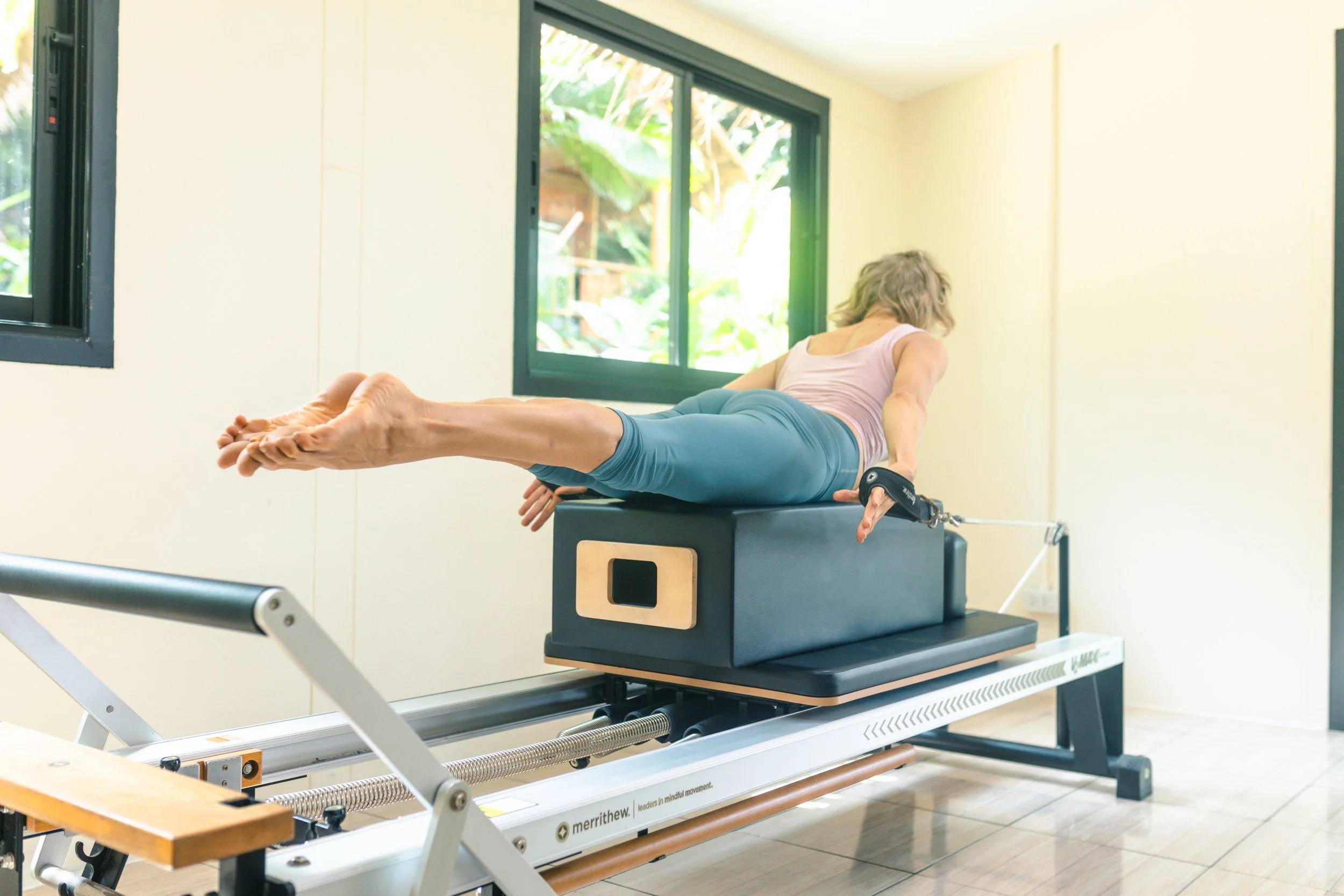 Woman performing Pilates exercise on a reformer machine in a bright room with large windows showing greenery outside.